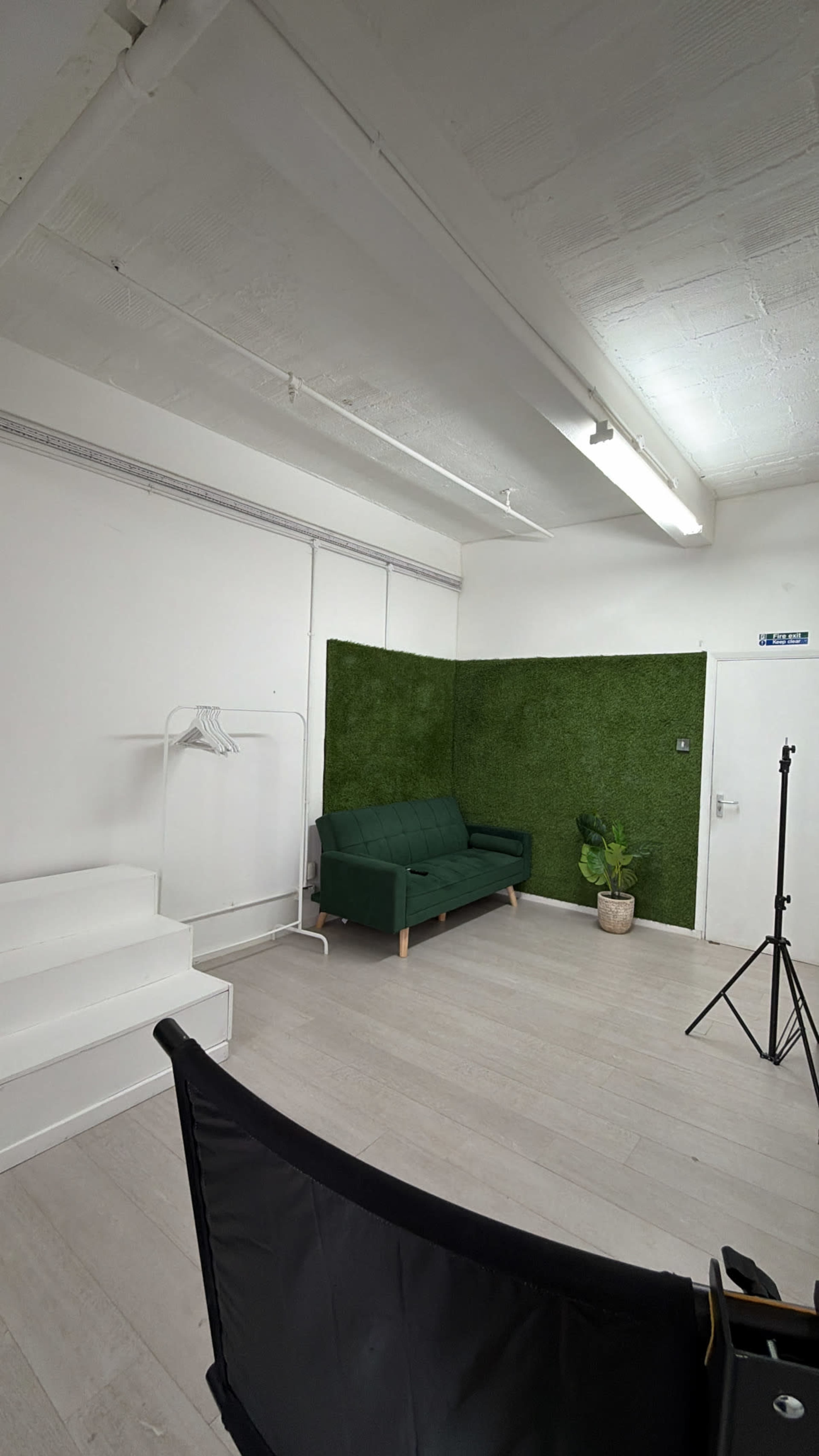 The image shows a minimalist studio space featuring a green couch, a decorative plant, and a backdrop of artificial grass against a white wall.