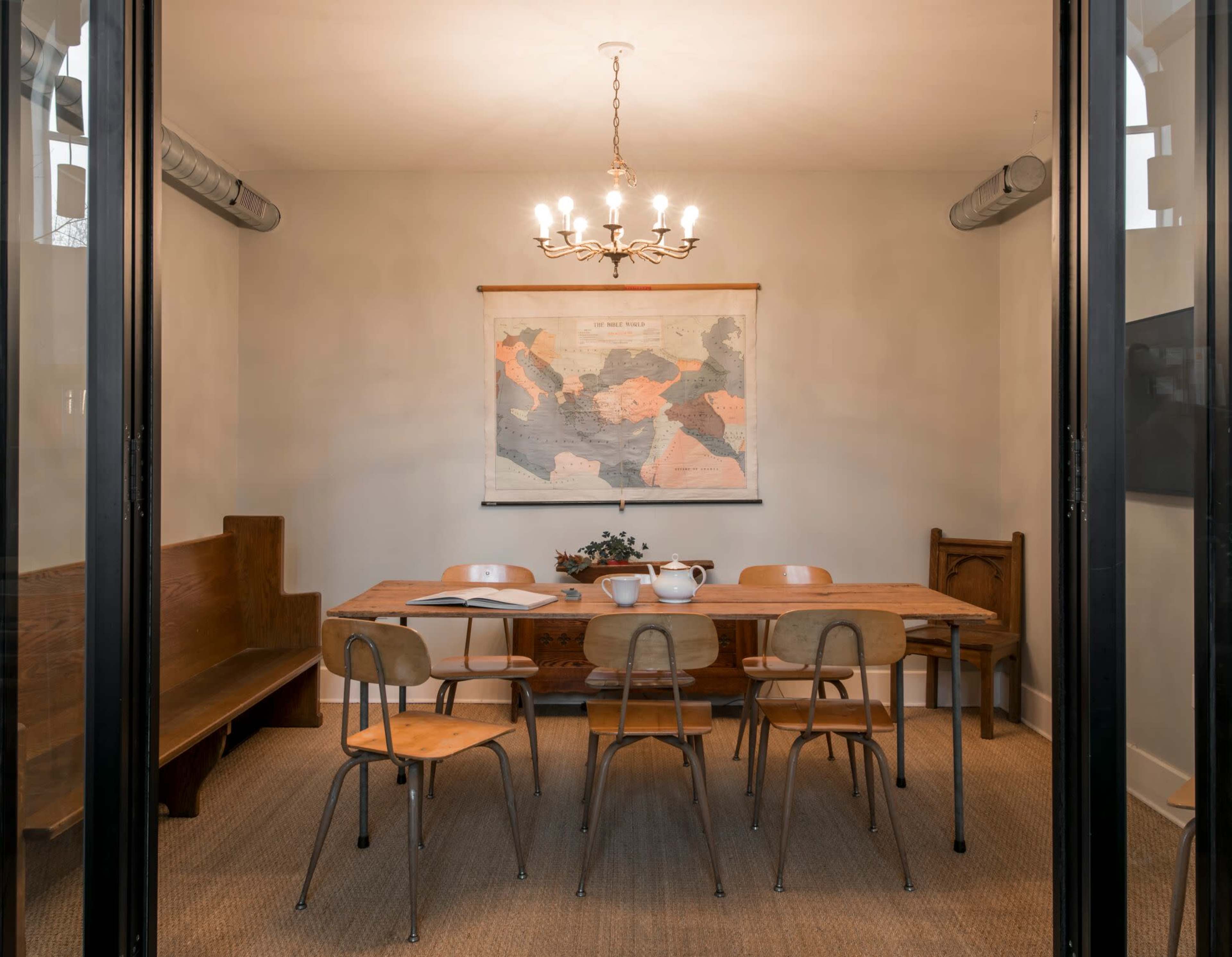 A spacious meeting room features a long wooden table surrounded by six chairs, with a large map hanging on the wall and a chandelier overhead.