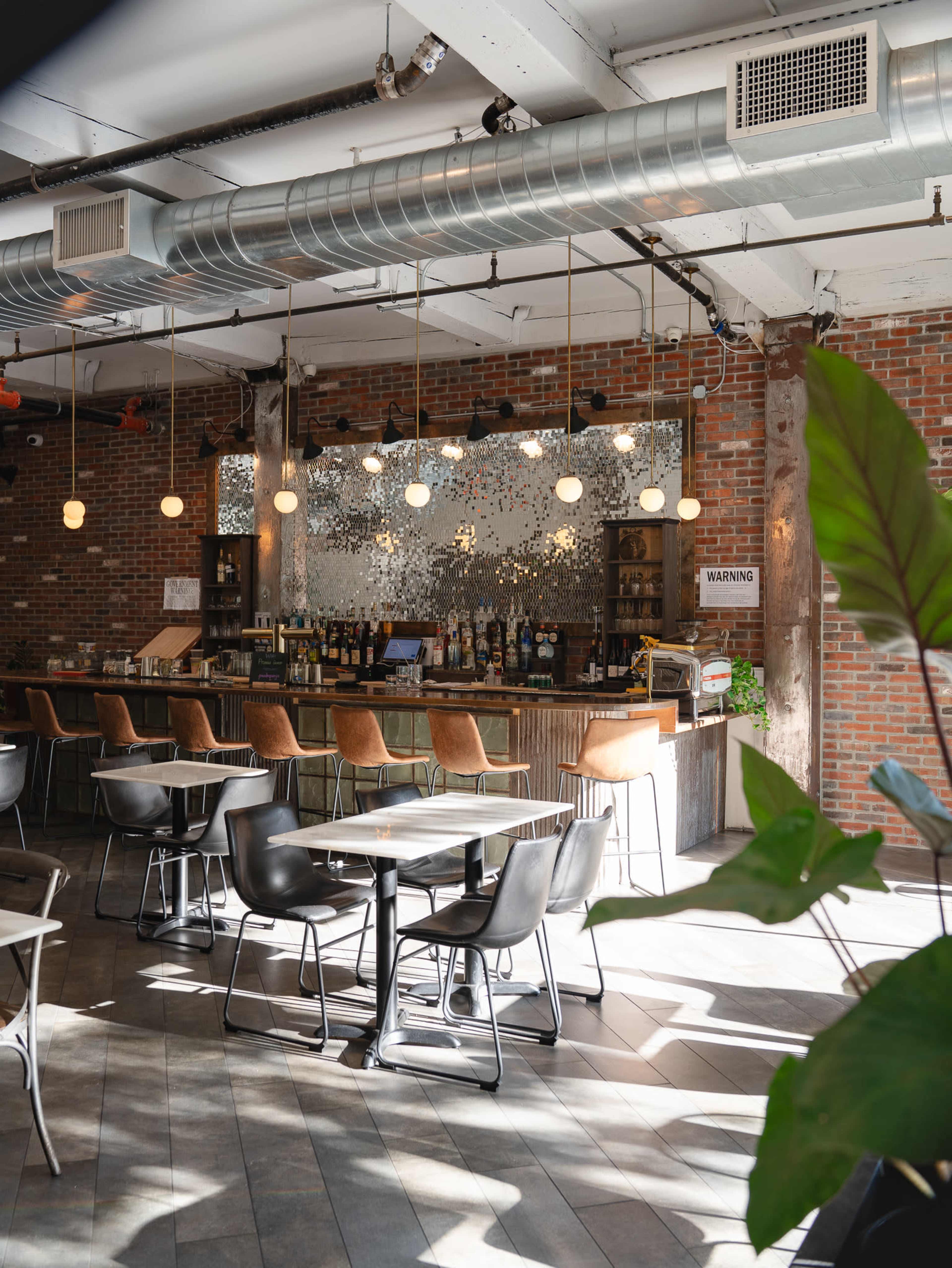 A modern café with brick walls, a bar area lined with bottles, and tables with chairs arranged around the space, illuminated by pendant lights.