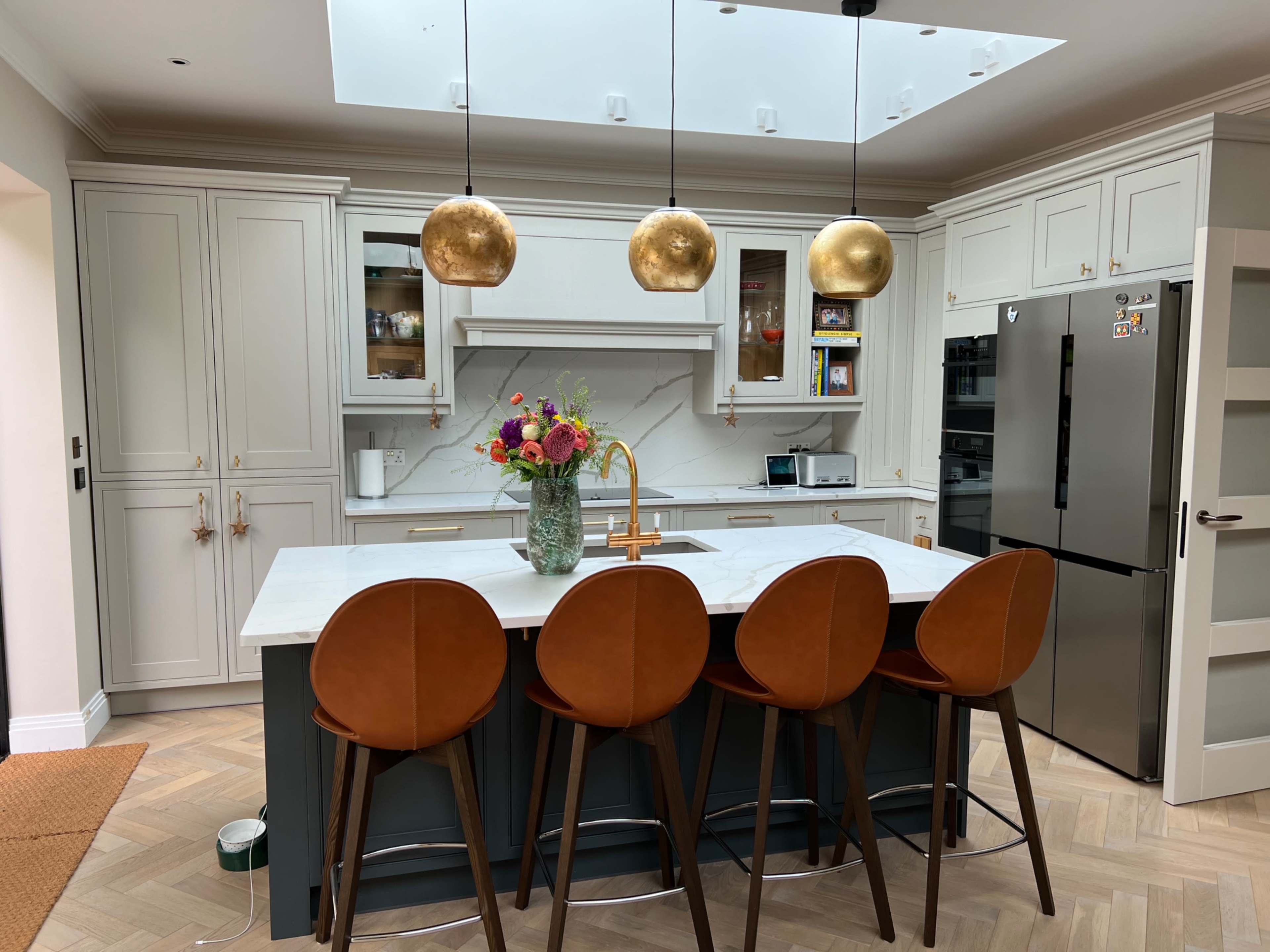 A modern kitchen features a large island with four tan bar stools, golden pendant lights above, and a stylish arrangement of flowers in a vase.