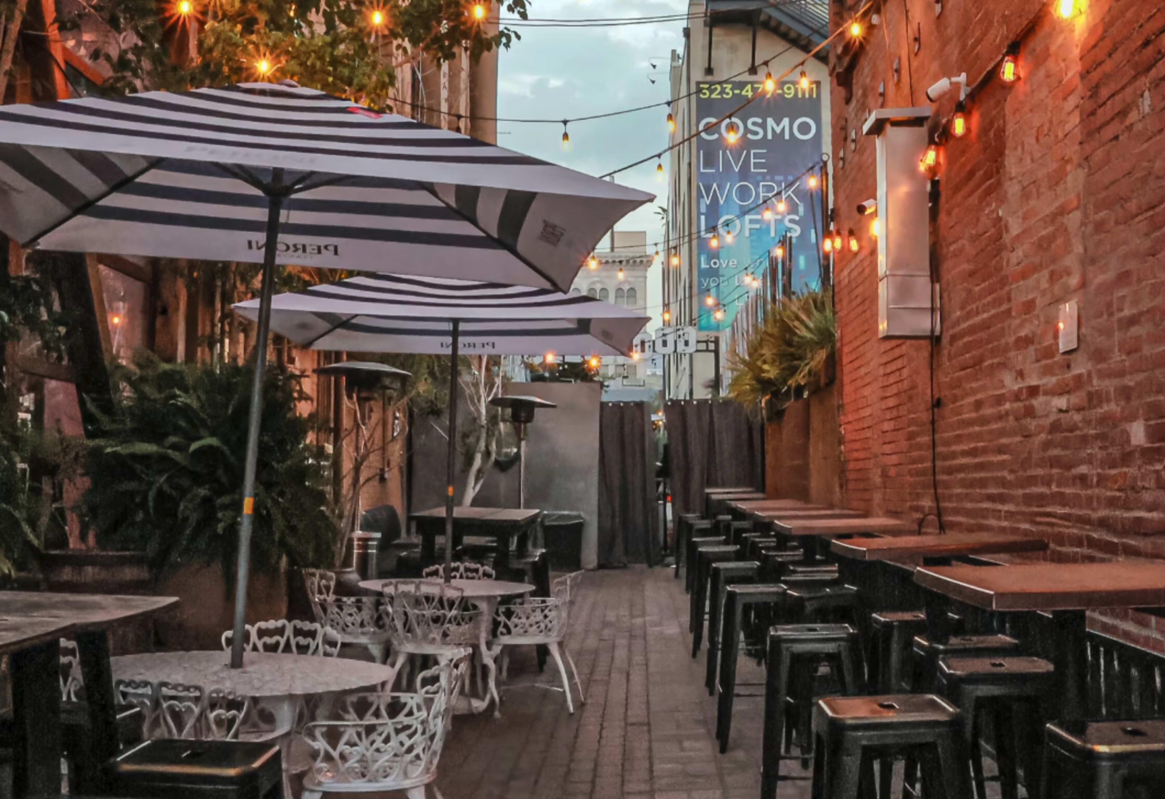 The image depicts a narrow outdoor alley lined with tables and chairs, illuminated by string lights and umbrellas, with a vibrant banner advertising "COSMO LIVE WORK LOFTS" in the background.