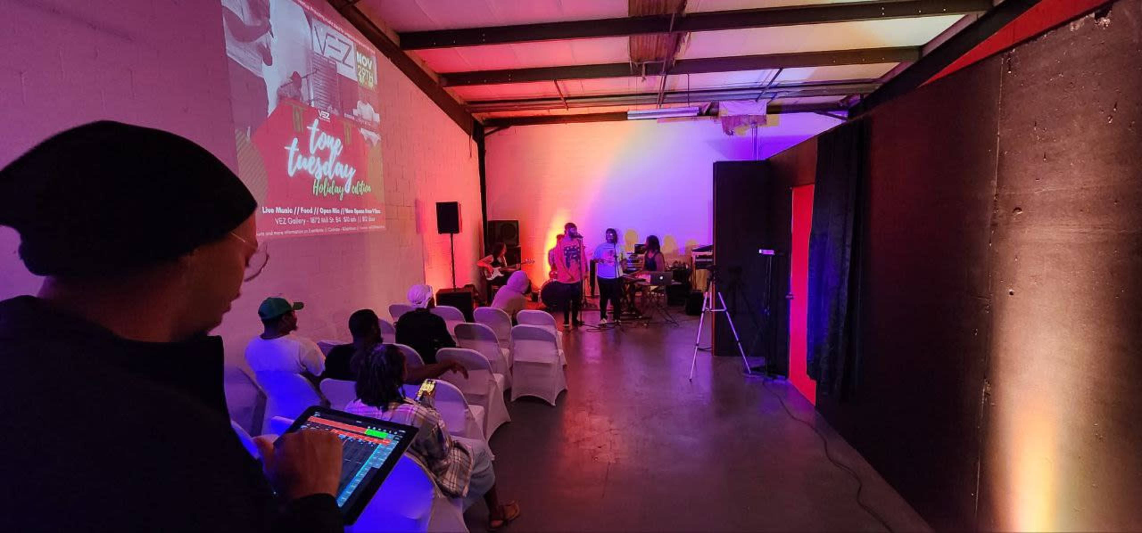 A small audience sits in a dimly lit room with white chairs, while performers engage on stage and a person controls sound equipment from the side.