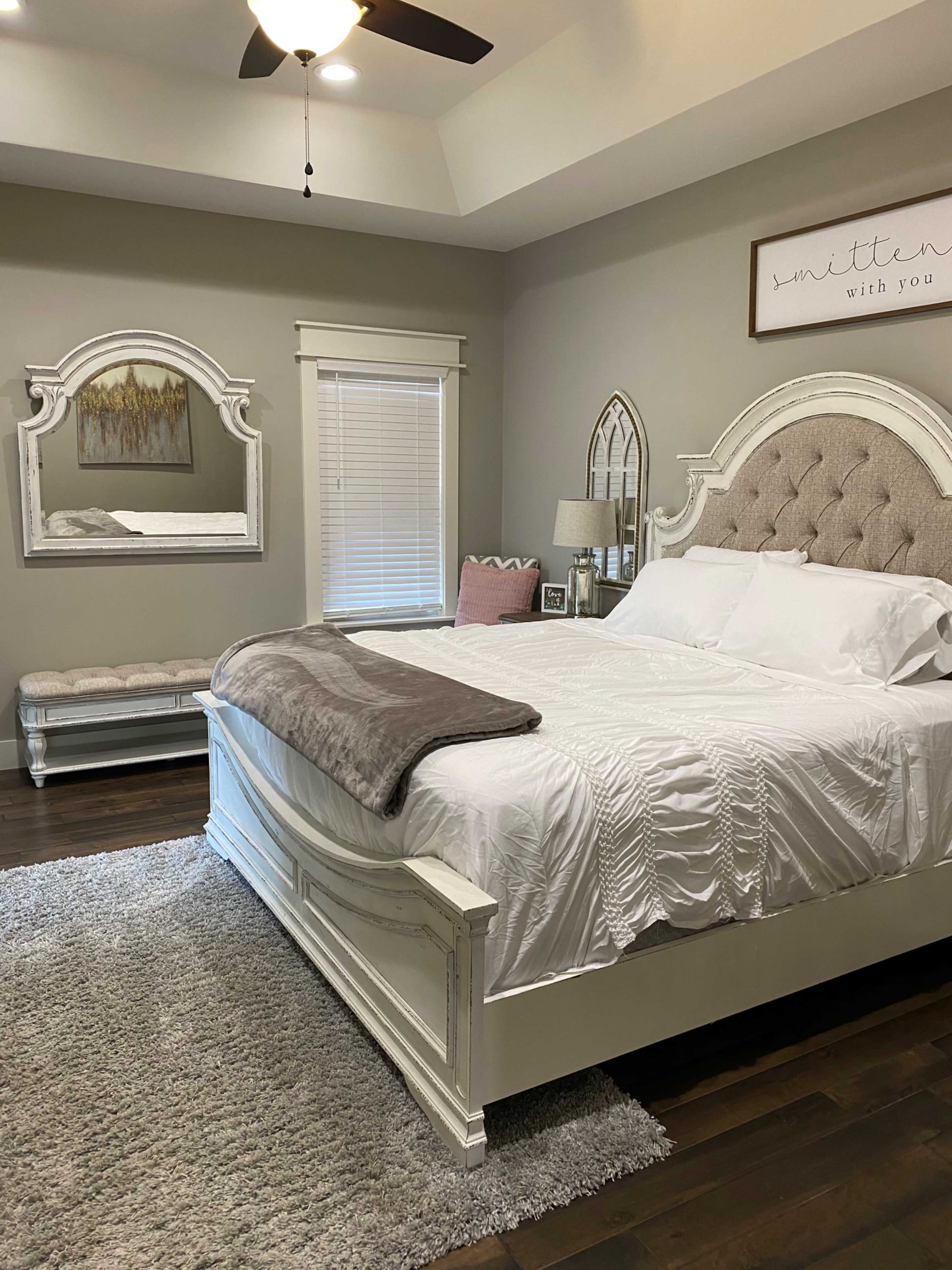 A neatly arranged bedroom featuring a large white bed with a tufted headboard, a gray throw blanket, a bench at the foot of the bed, a mirror, and a window with blinds.