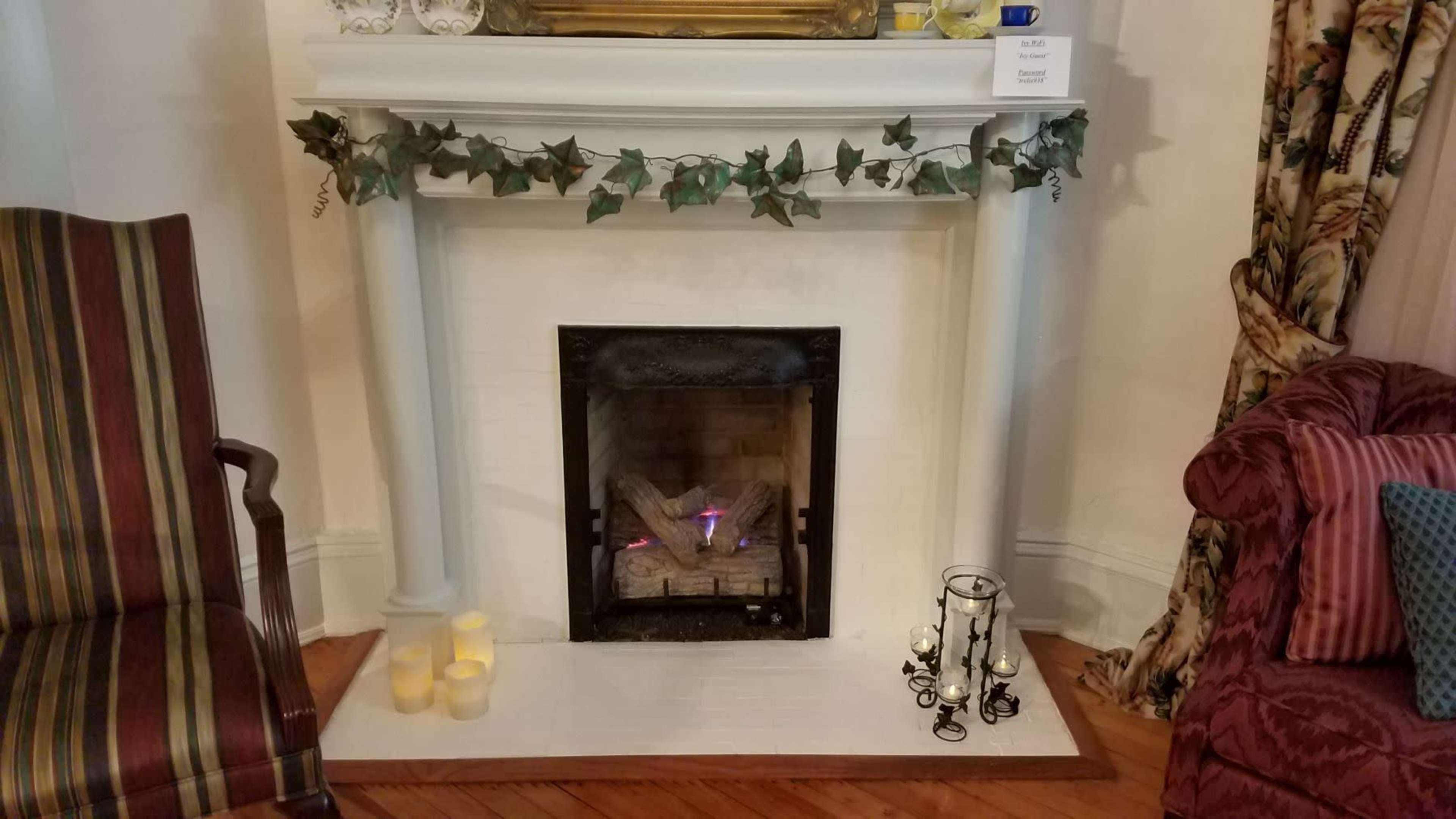 The image shows a decorative fireplace with an electric log set, surrounded by candles and adorned with ivy, situated between two upholstered chairs.