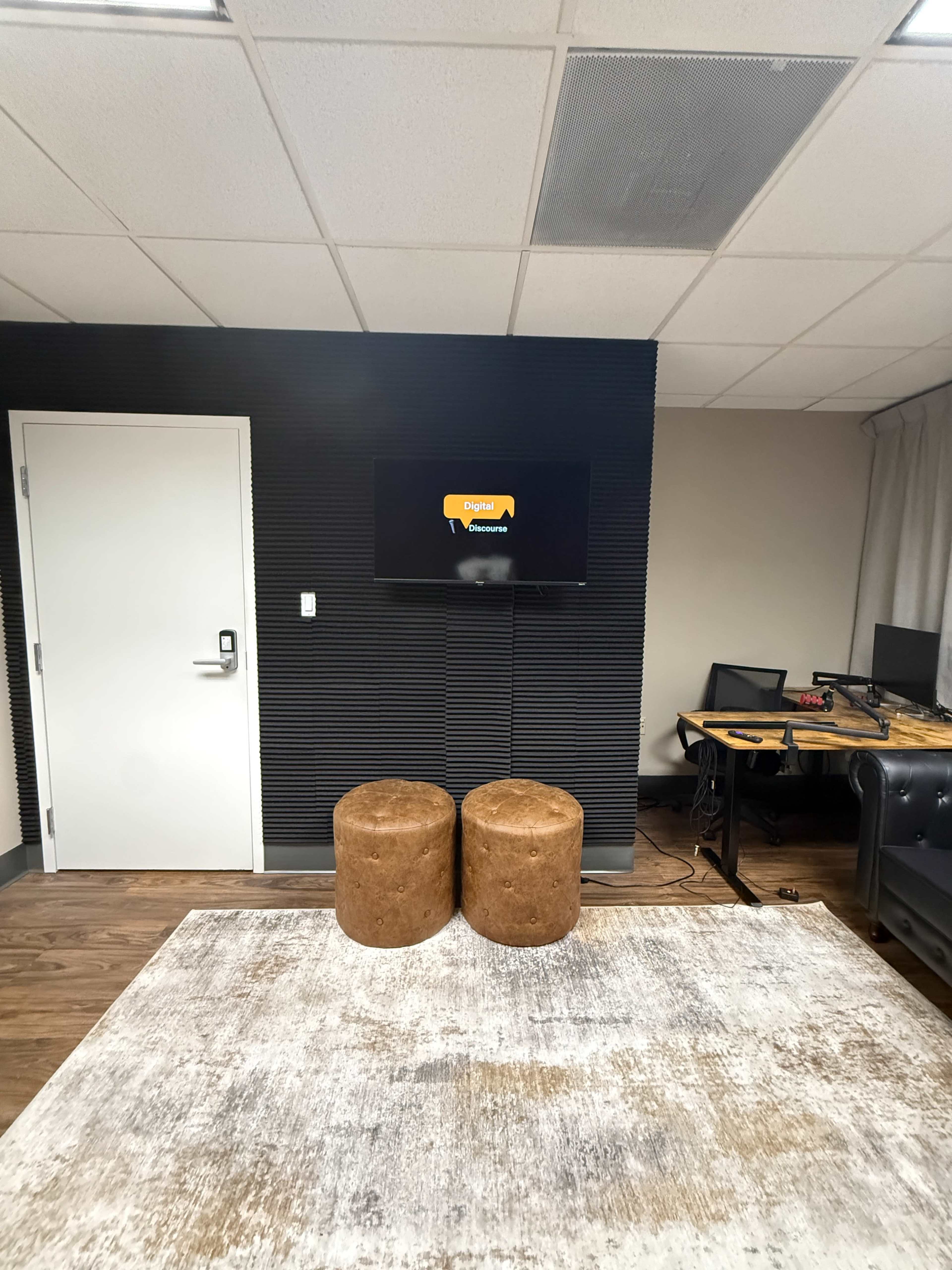 The image shows a modern office space featuring two round brown stools in front of a wall-mounted television, with a minimalist desk setup and a beige rug on the floor.