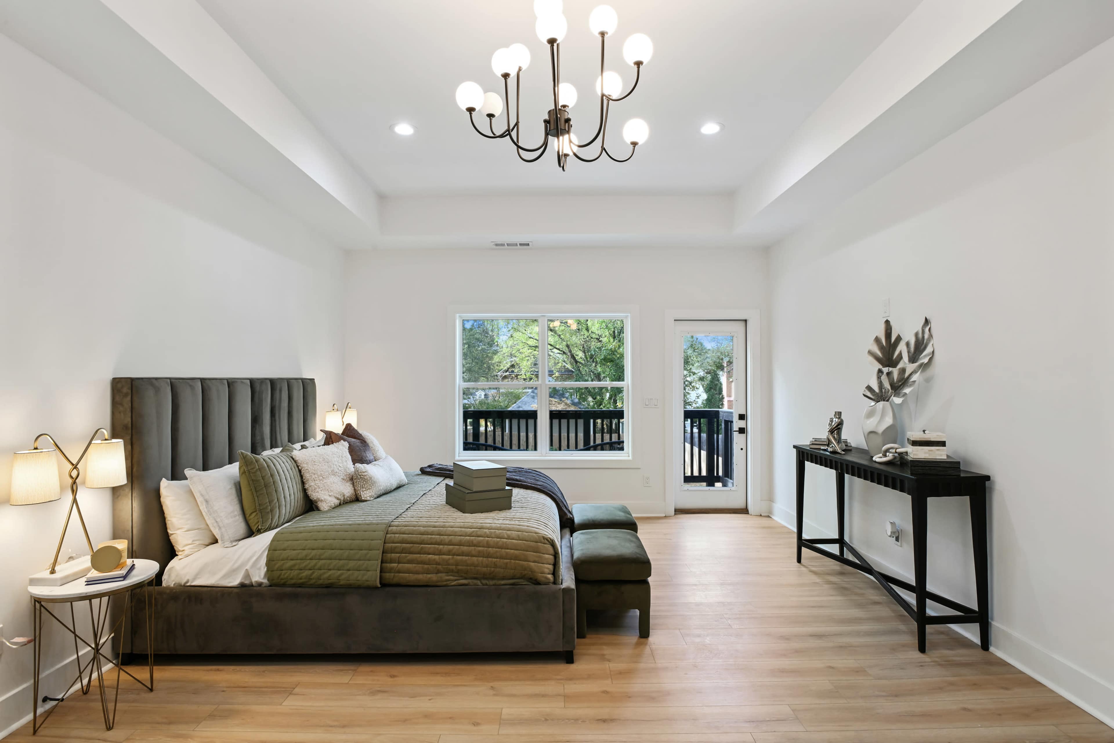 The image shows a spacious bedroom featuring a large bed with a tufted headboard, a dark wood side table, and a minimalist decorative console along one wall.