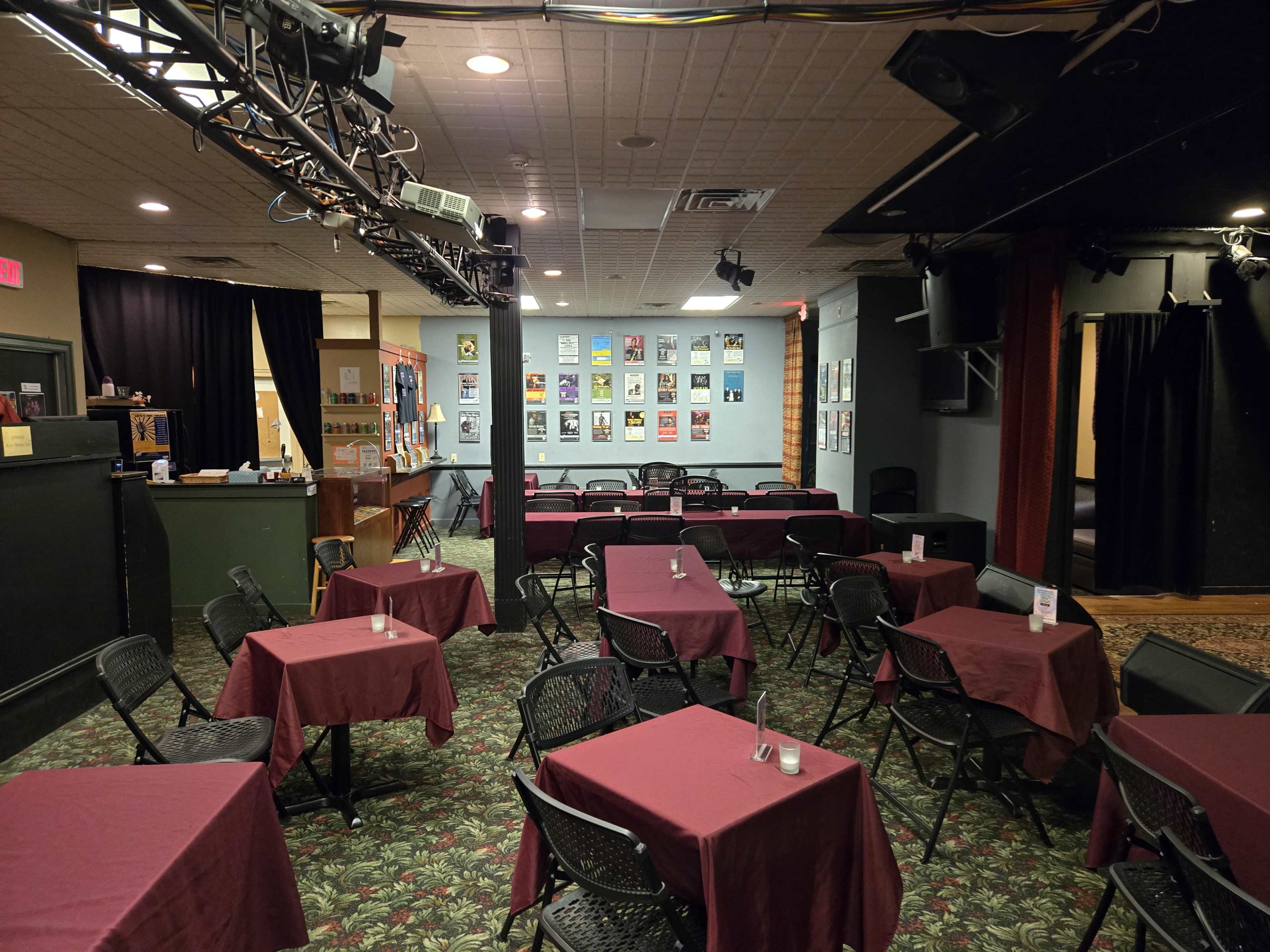 The image shows a small performance venue with rows of tables covered in burgundy cloth and a stage area in the background featuring posters on the walls.