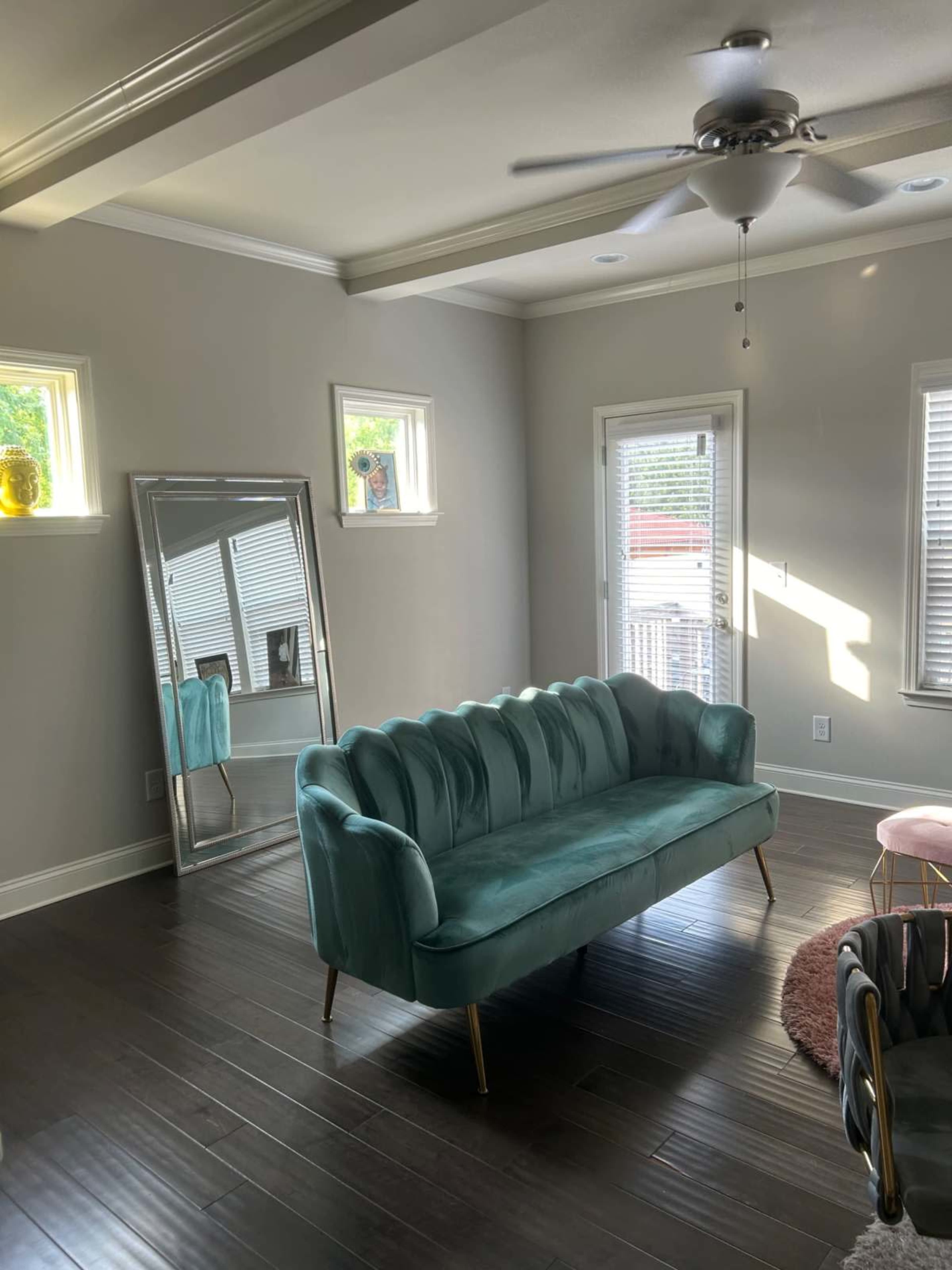 The room features a turquoise couch with gold legs, a large mirror, and multiple windows allowing natural light to enter.