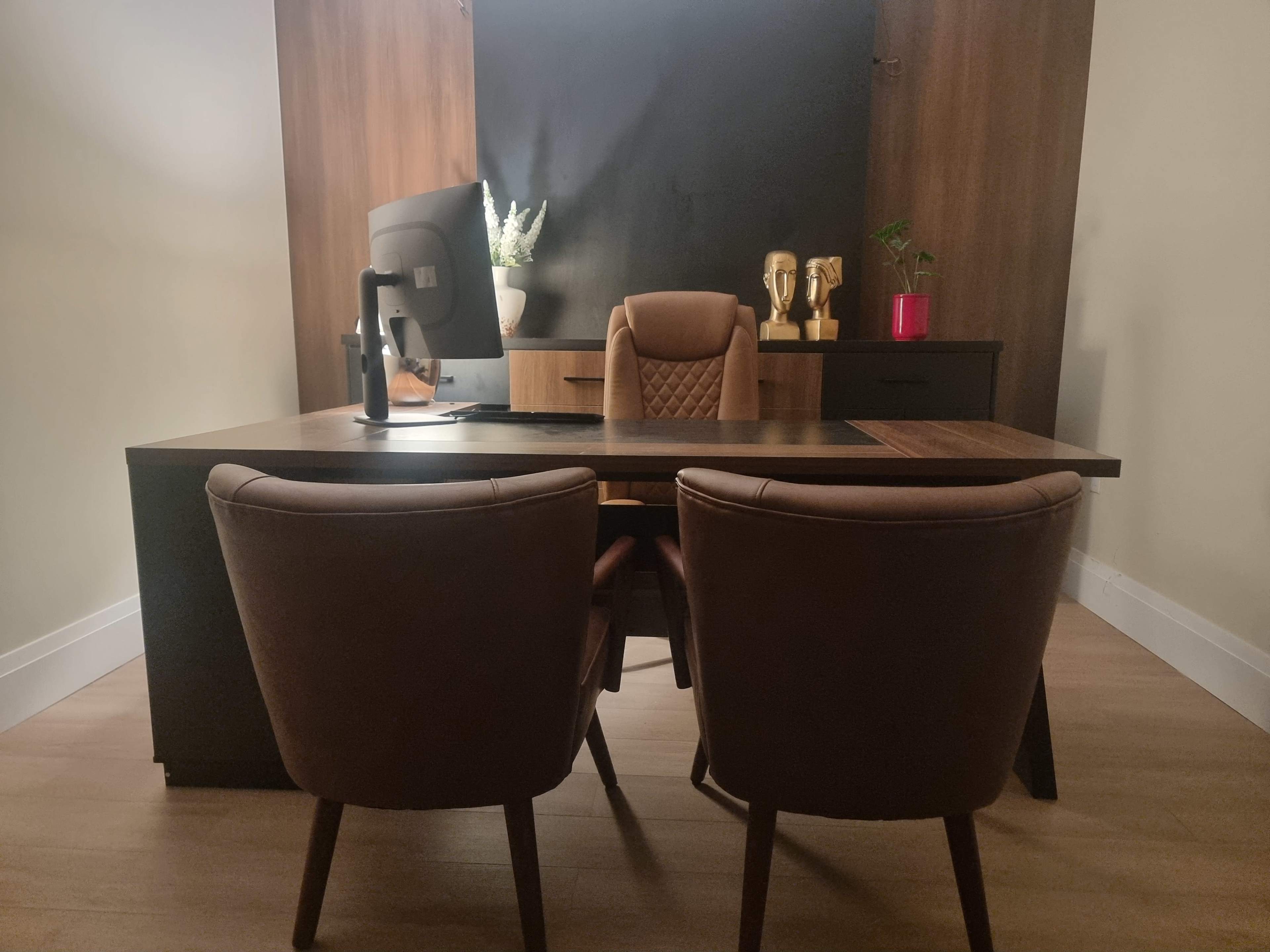 The image shows a modern office space featuring a dark wood desk, two brown chairs, a computer monitor, and a potted plant on a shelf.