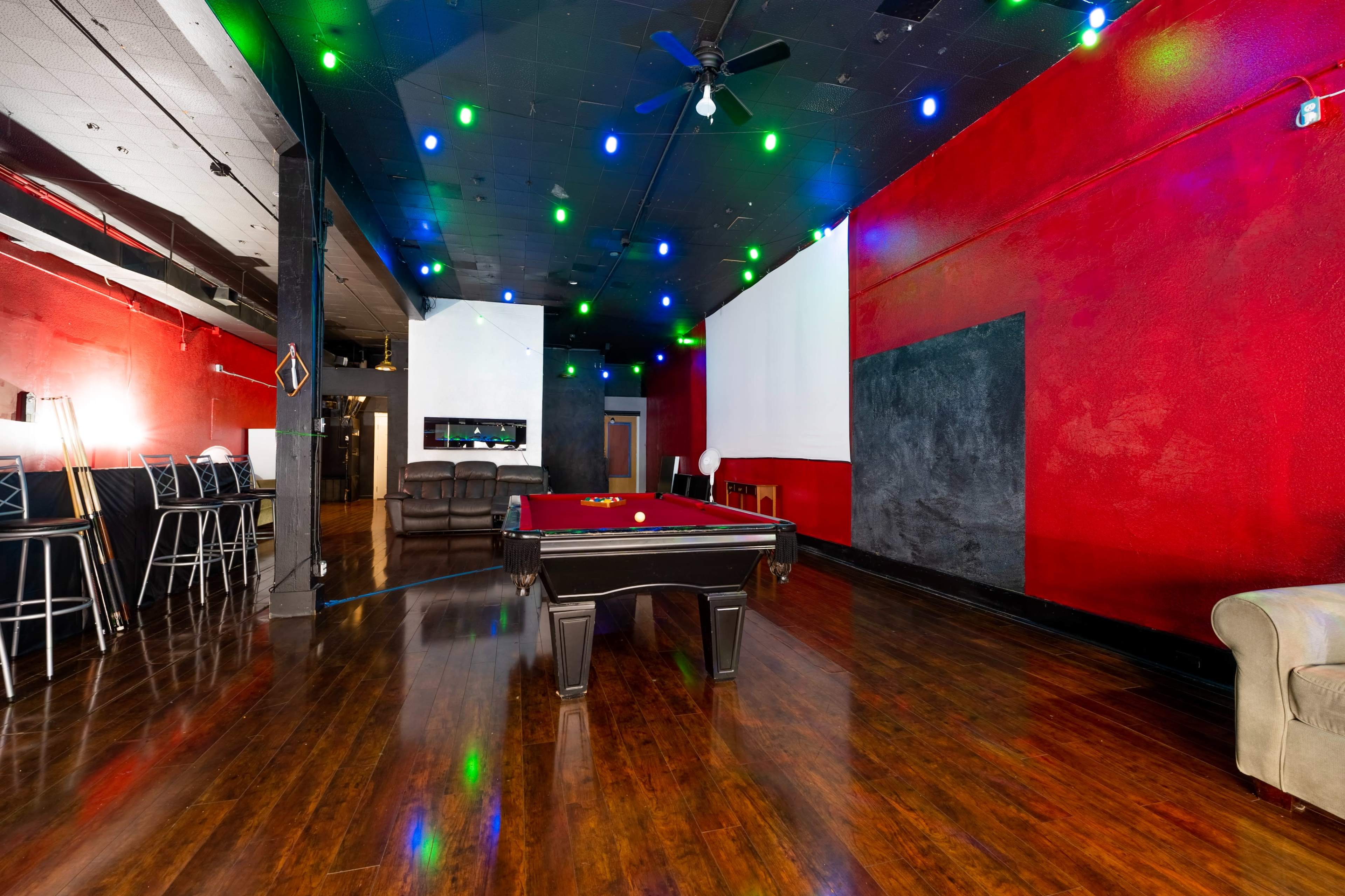 A game room with a billiard table in the center, surrounded by red walls, hardwood floors, and colorful ceiling lights.