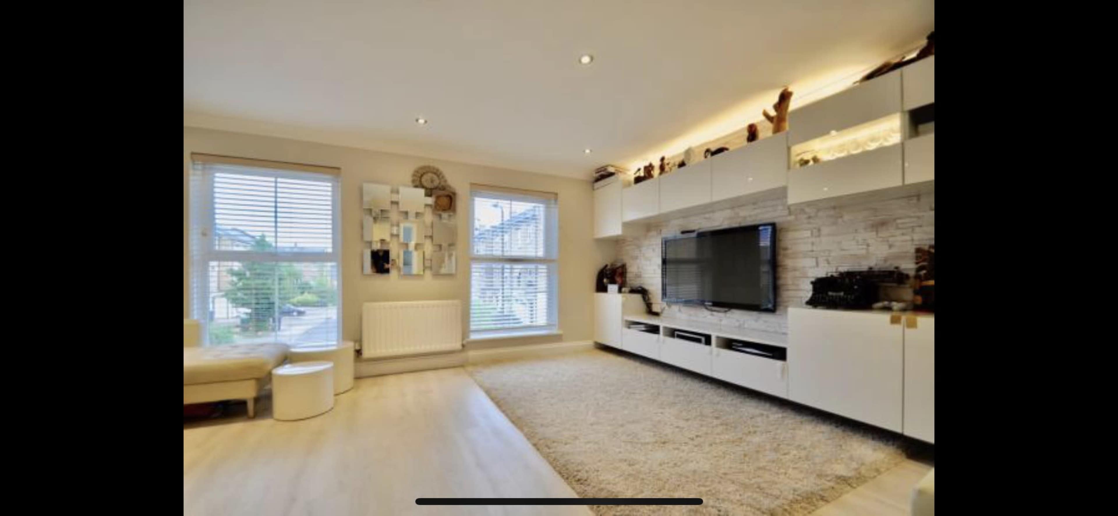 A modern living room with a television mounted on the wall, a plush rug on the floor, and a large window with blinds.