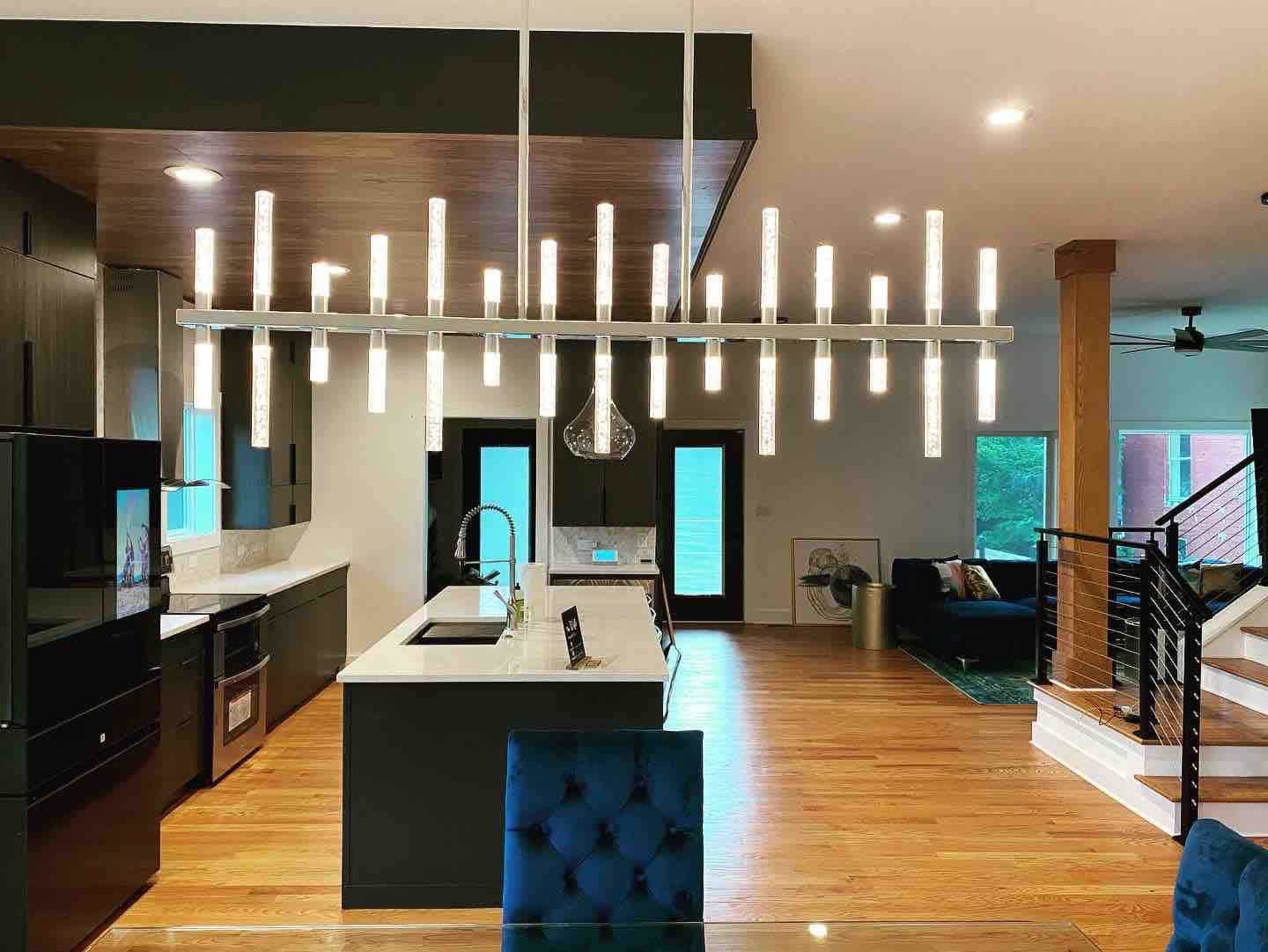 A modern kitchen and dining area featuring a long, sleek light fixture above a dining table, with dark cabinetry and wooden floors.