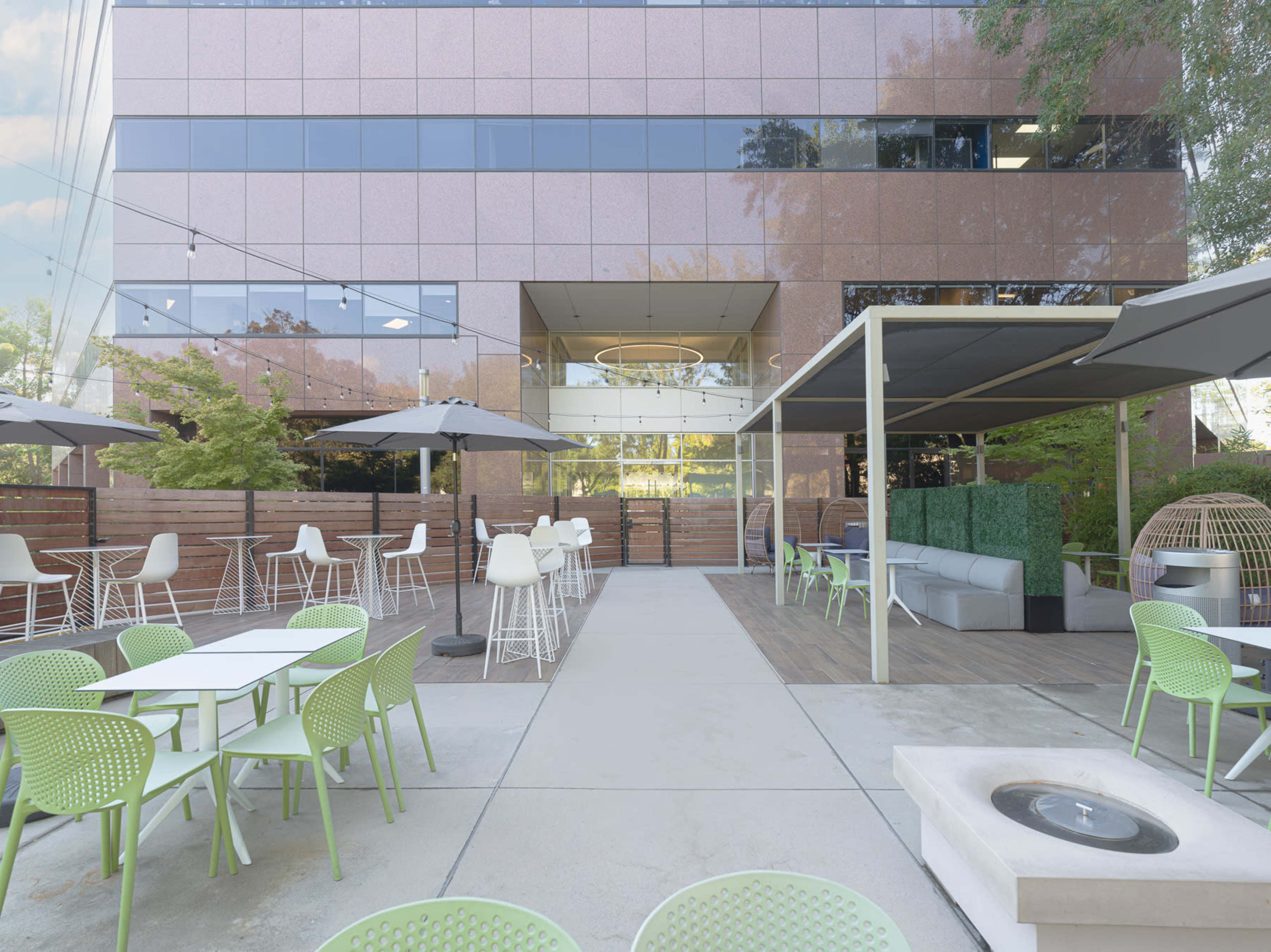 The image shows an outdoor patio area with white tables and green chairs, surrounded by modern architecture and landscaping, featuring umbrellas and a fire pit.