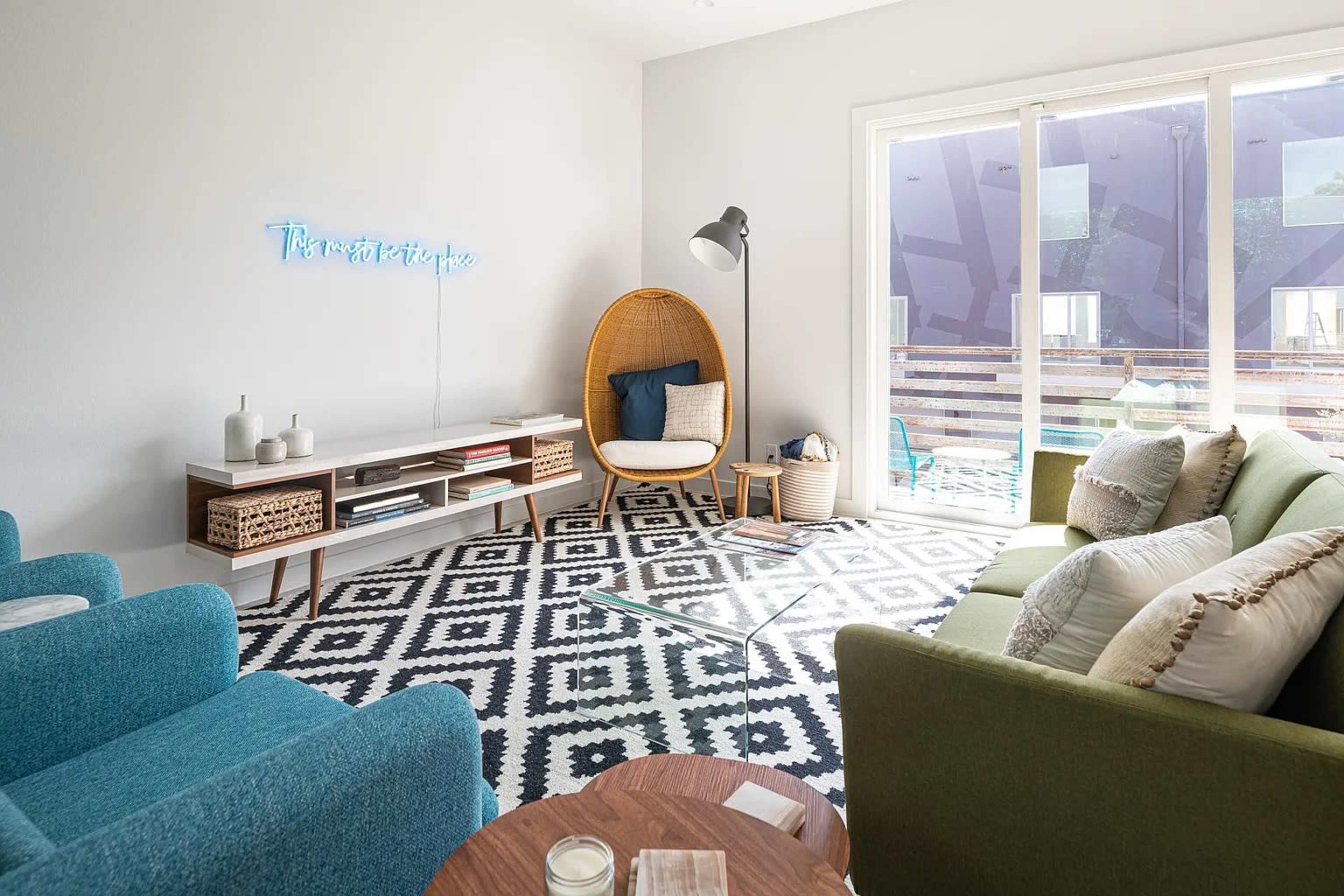 A modern living room with teal and green chairs, a decorative rug, a large window, and a rattan chair beside a side table.