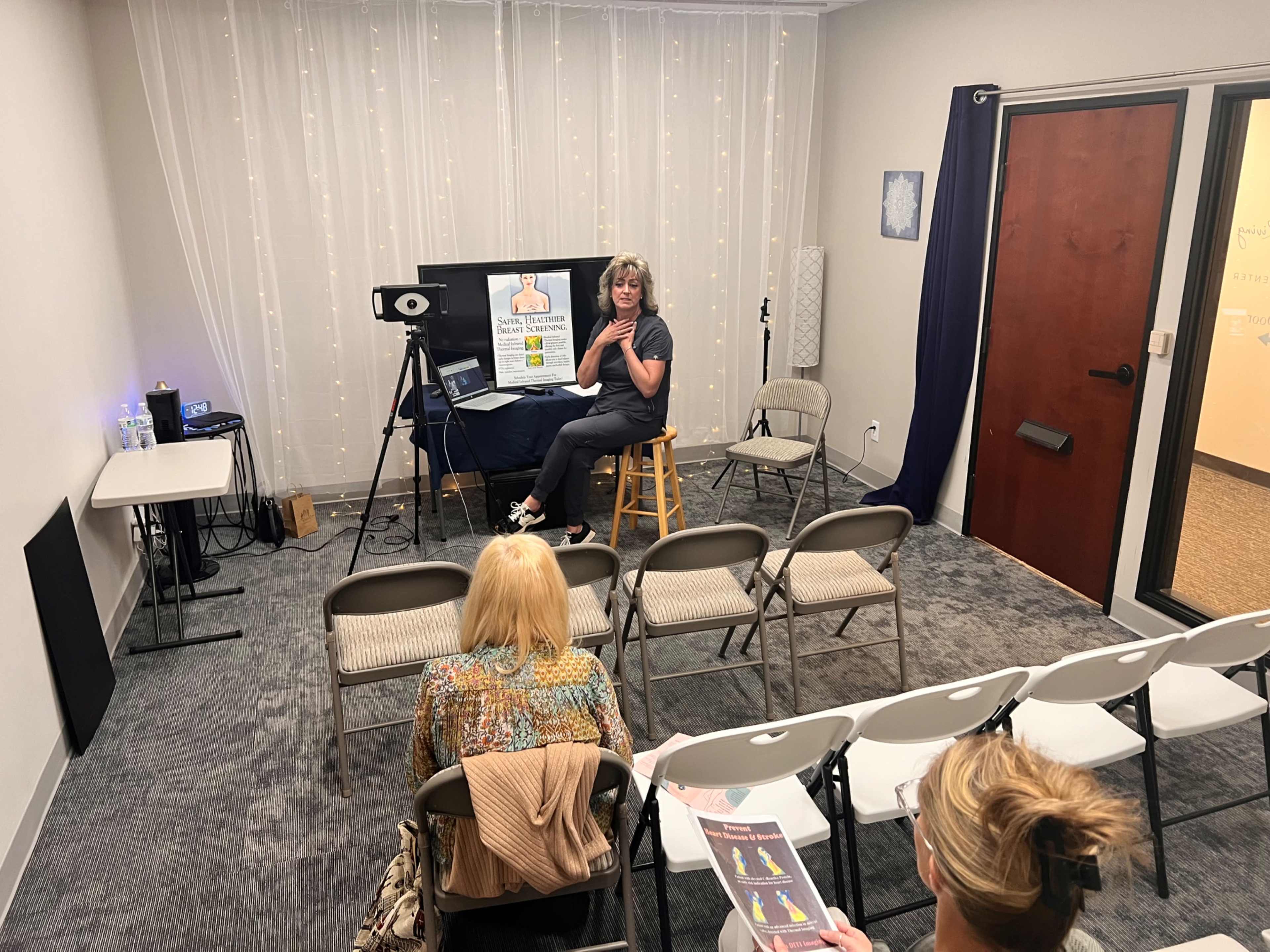 A woman is seated on a stool in front of a projector screen while speaking to an audience in a small room with folding chairs.