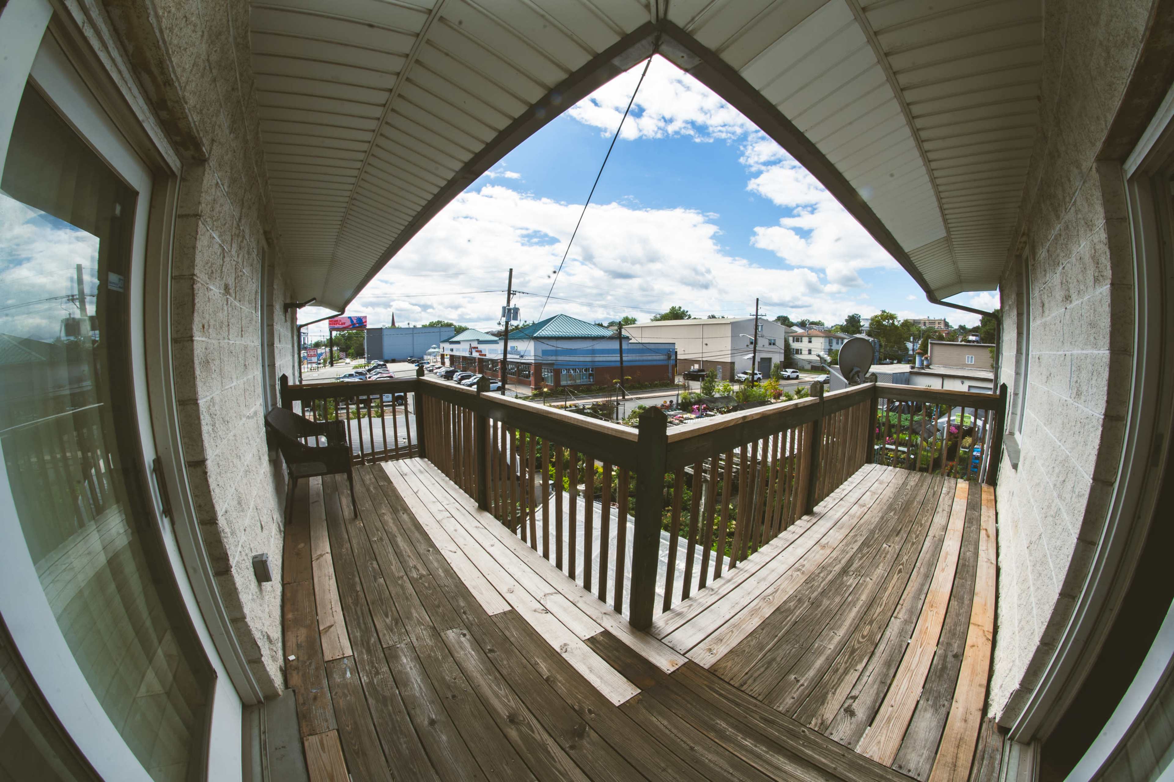 The image shows a wooden balcony with a view of a town street and buildings under a partly cloudy sky.