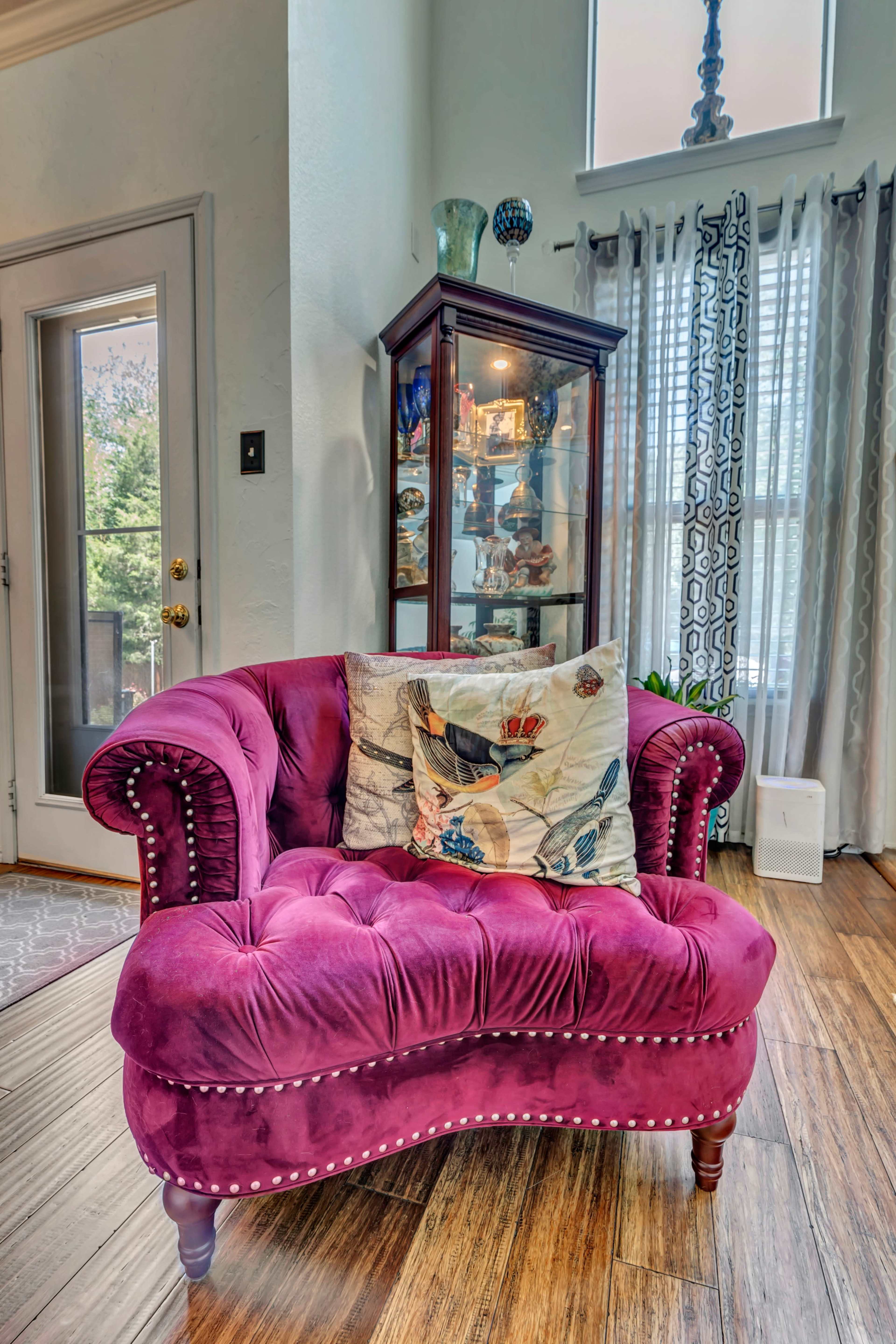 A purple velvet loveseat with decorative pillows is positioned near a glass display cabinet and large windows in a bright room.