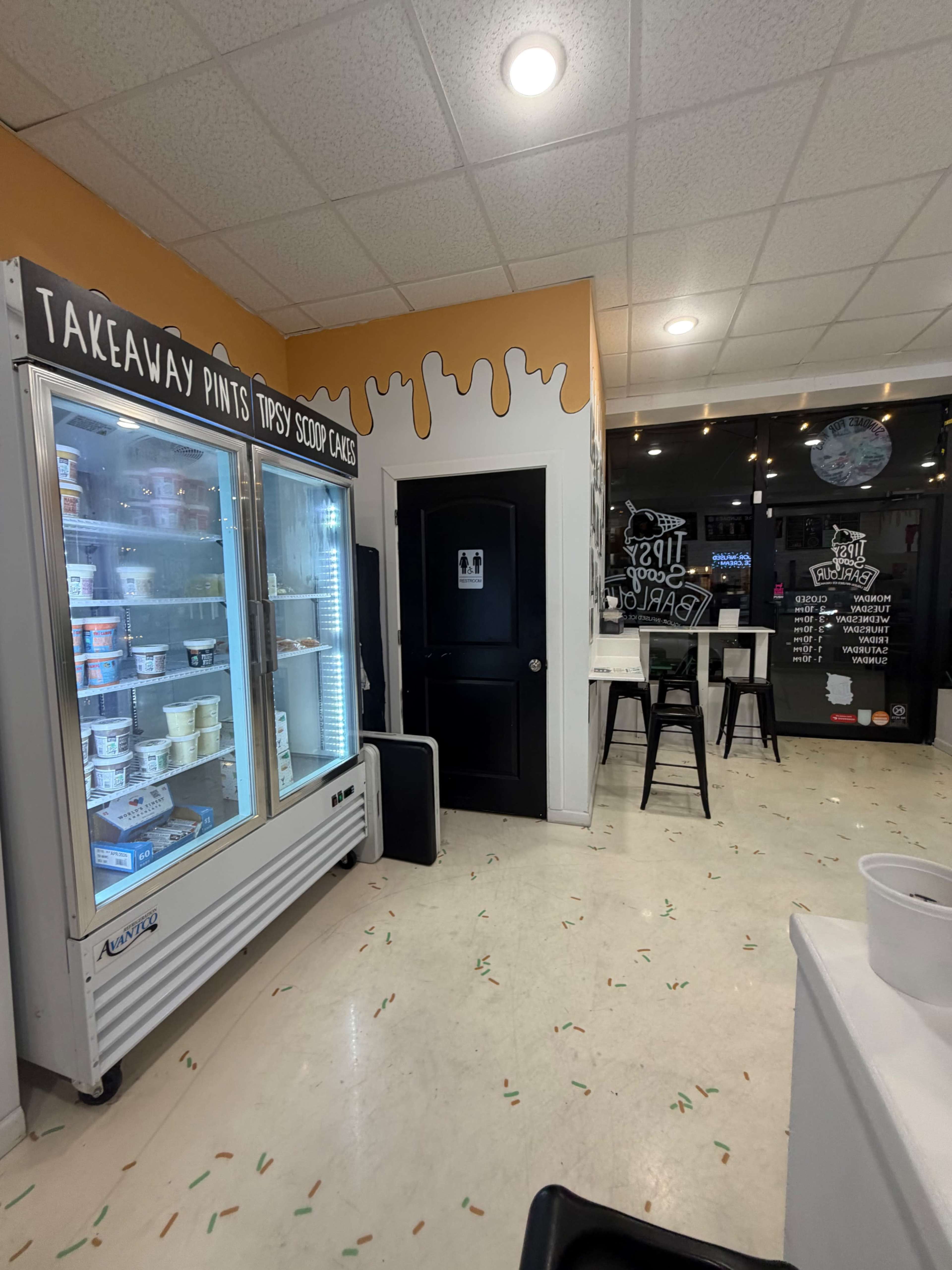The image displays a brightly lit ice cream shop interior featuring a glass freezer filled with ice cream containers, a black restroom door, and a seating area with high stools.