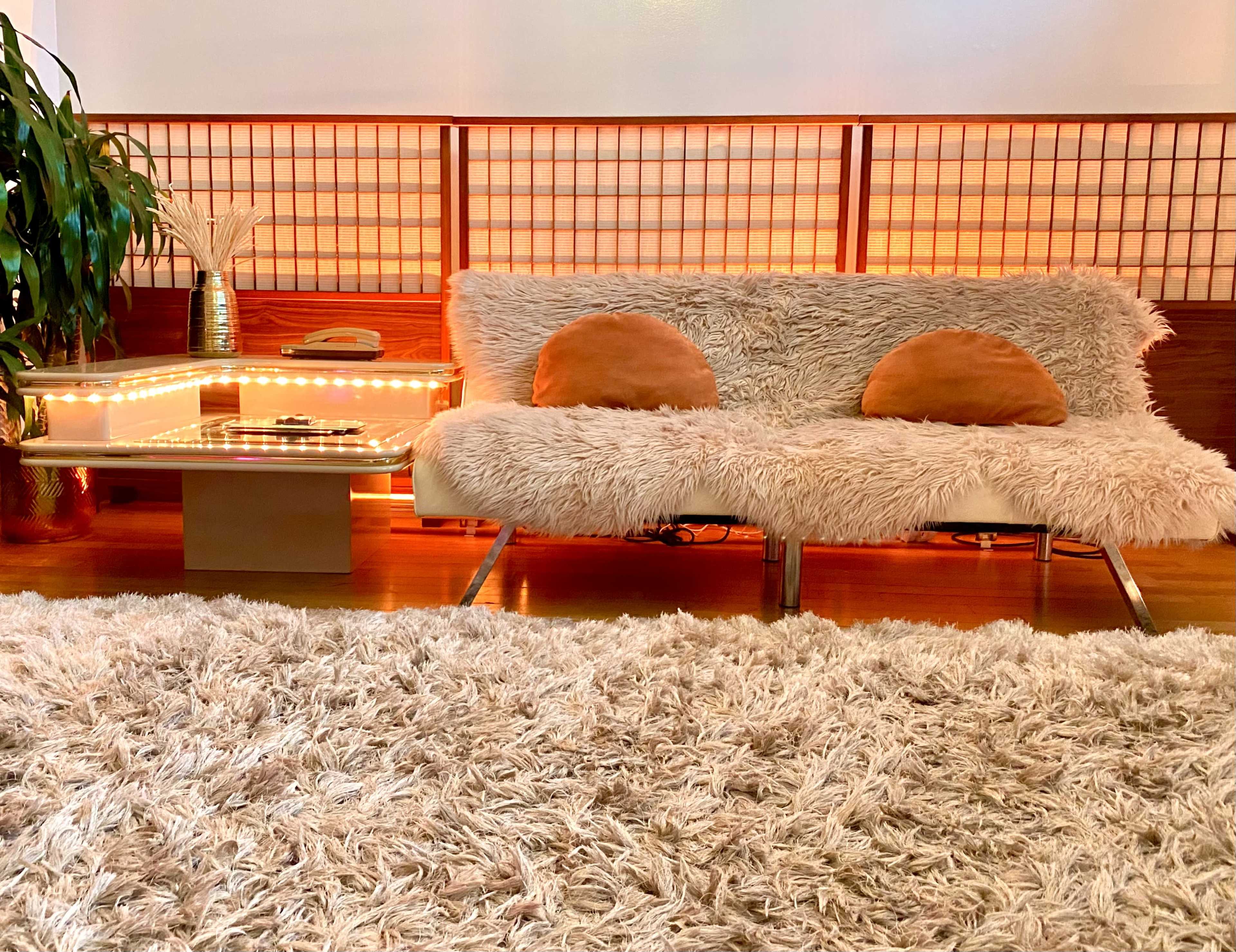 A cozy living room features a fluffy white sofa with round orange cushions, a lighted wooden side table, and a thick, shaggy rug on the wooden floor.