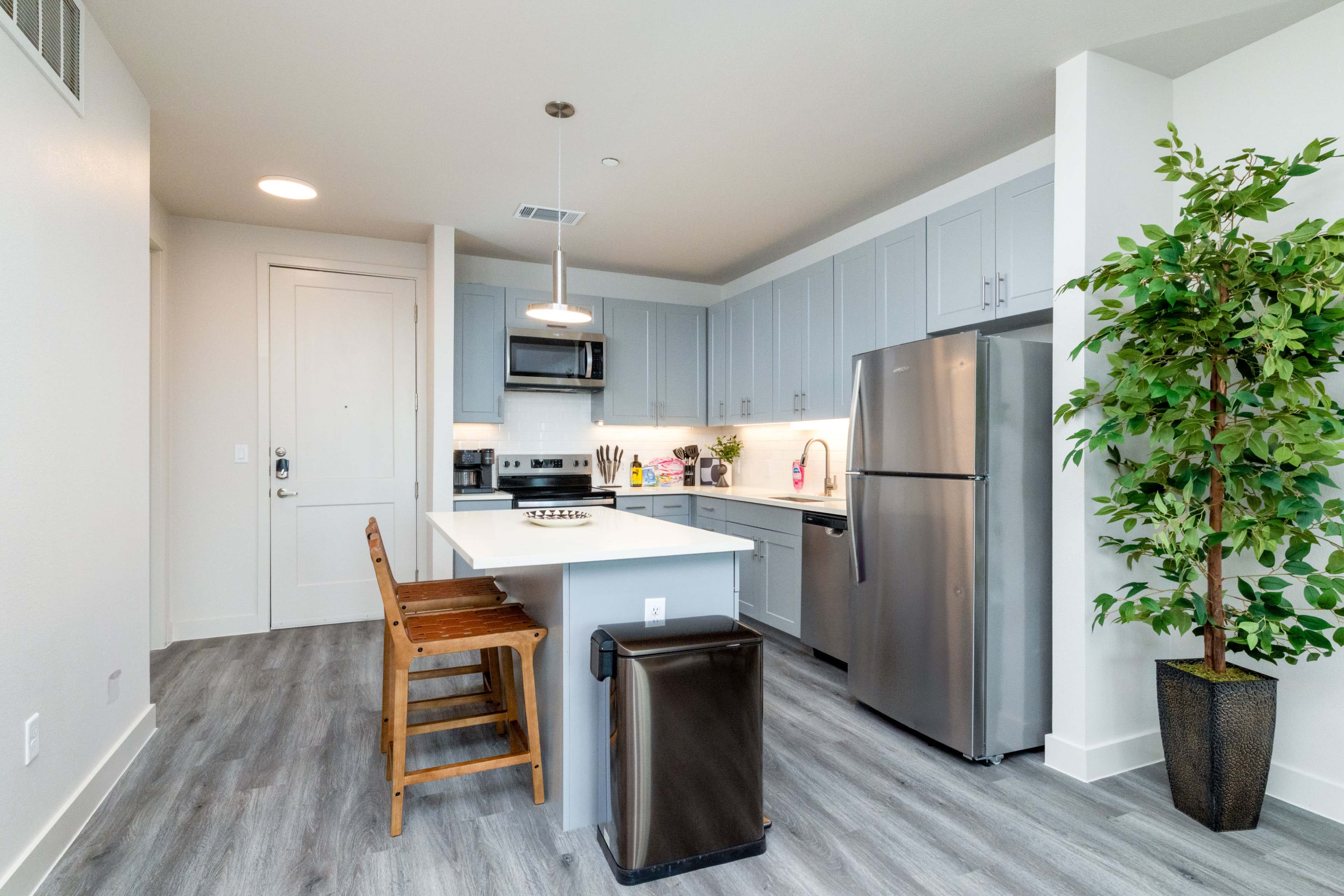 The image shows a modern kitchen with light gray cabinets, stainless steel appliances, a central island with seating, and a potted plant in the corner.