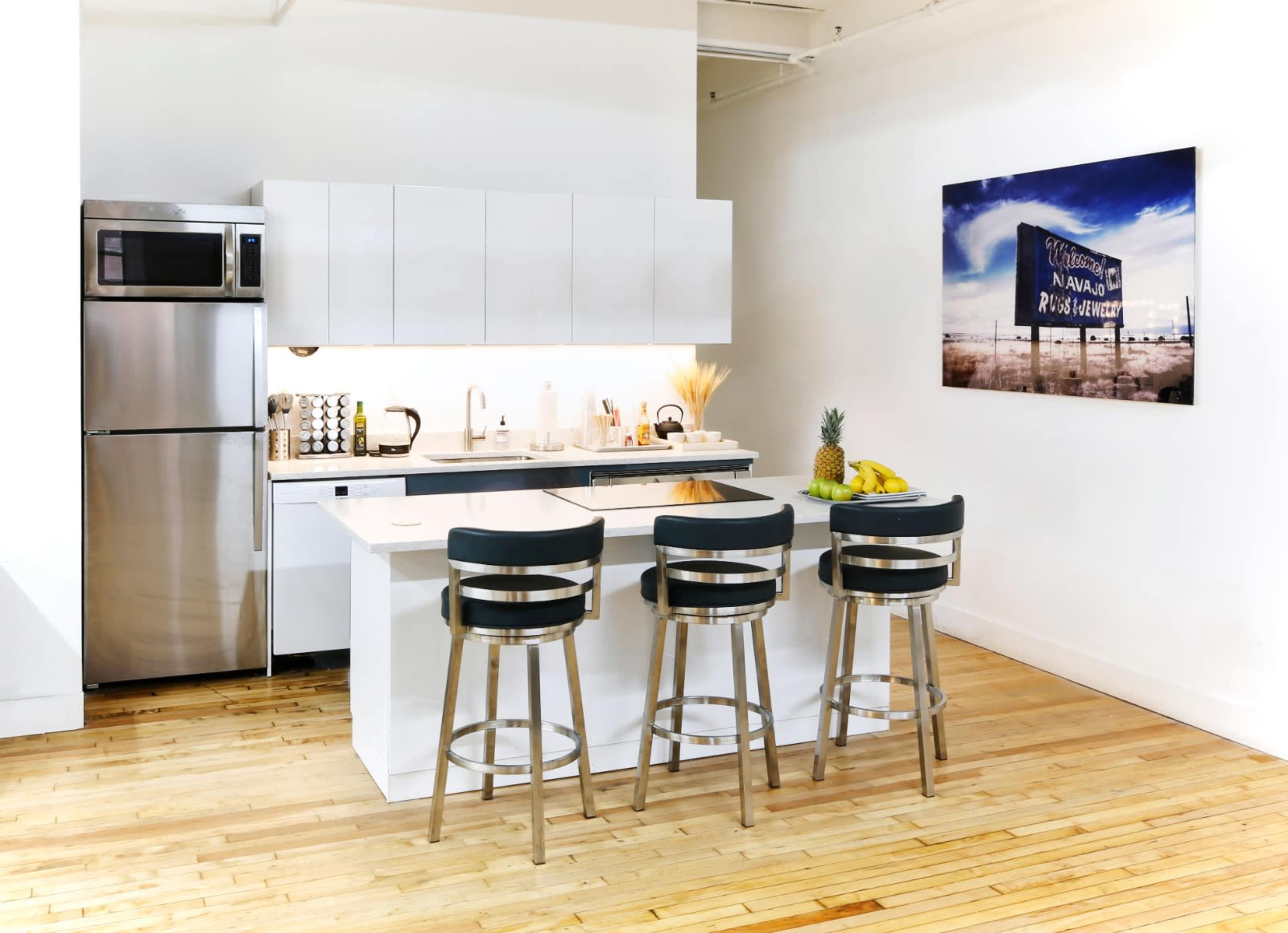 The image depicts a modern kitchen featuring a stainless steel refrigerator, white cabinetry, a countertop with seating for three, and a piece of wall art in the background.