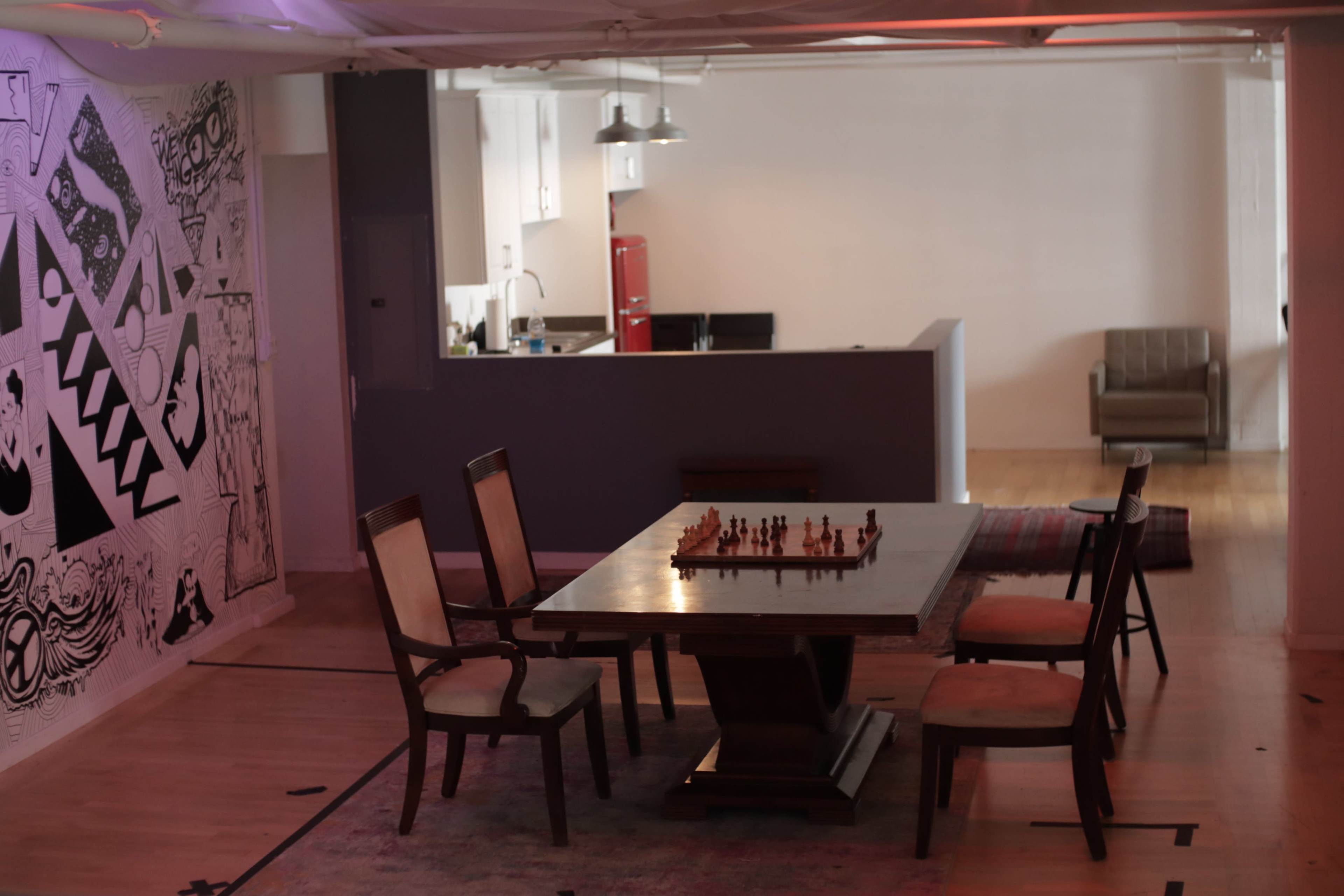 A wooden chess table surrounded by six chairs sits in a spacious, minimally furnished room with a kitchenette in the background.