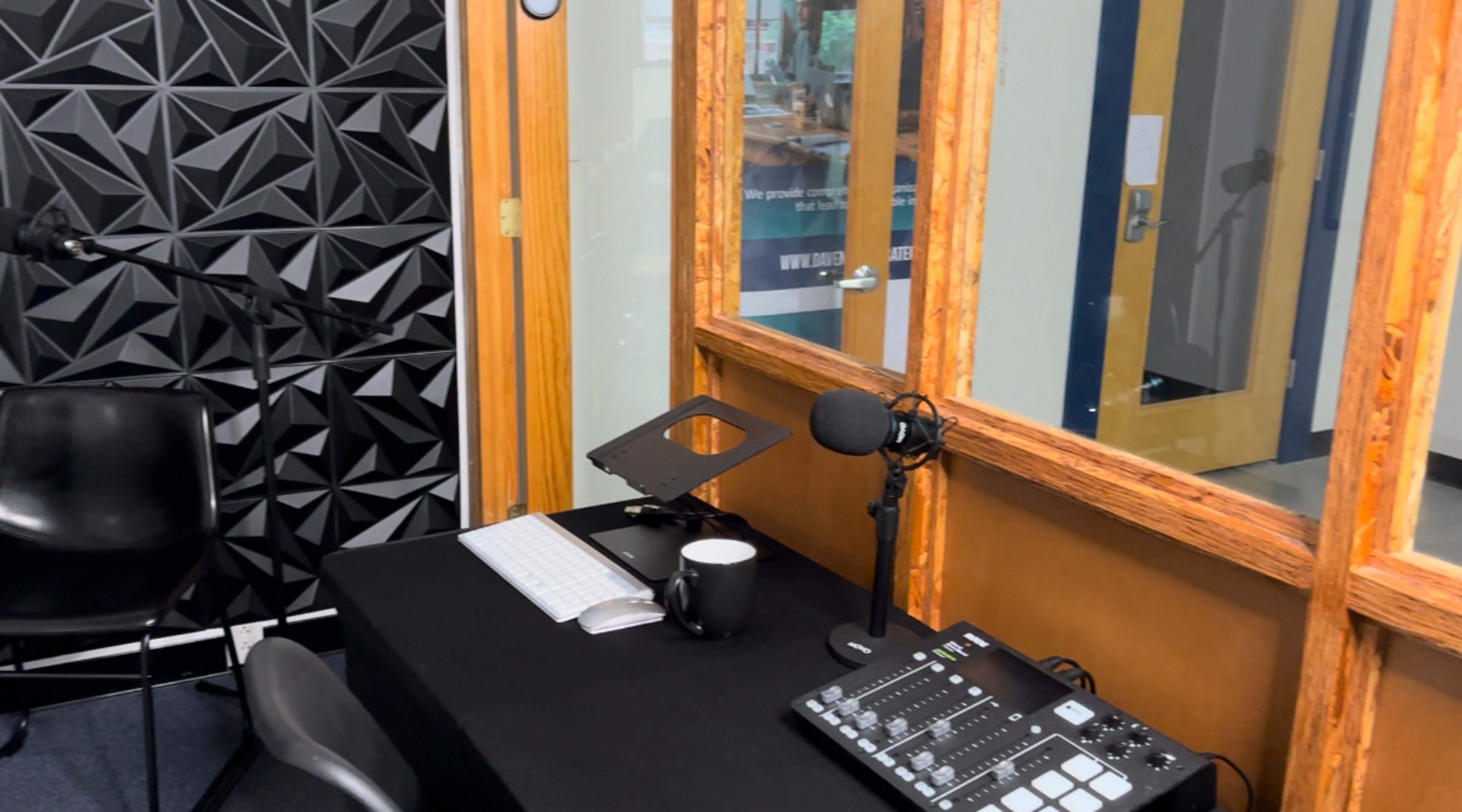A recording studio setup with a microphone, a control panel, a computer keyboard, and a coffee cup on a table, all within a room featuring soundproofing panels on the walls.