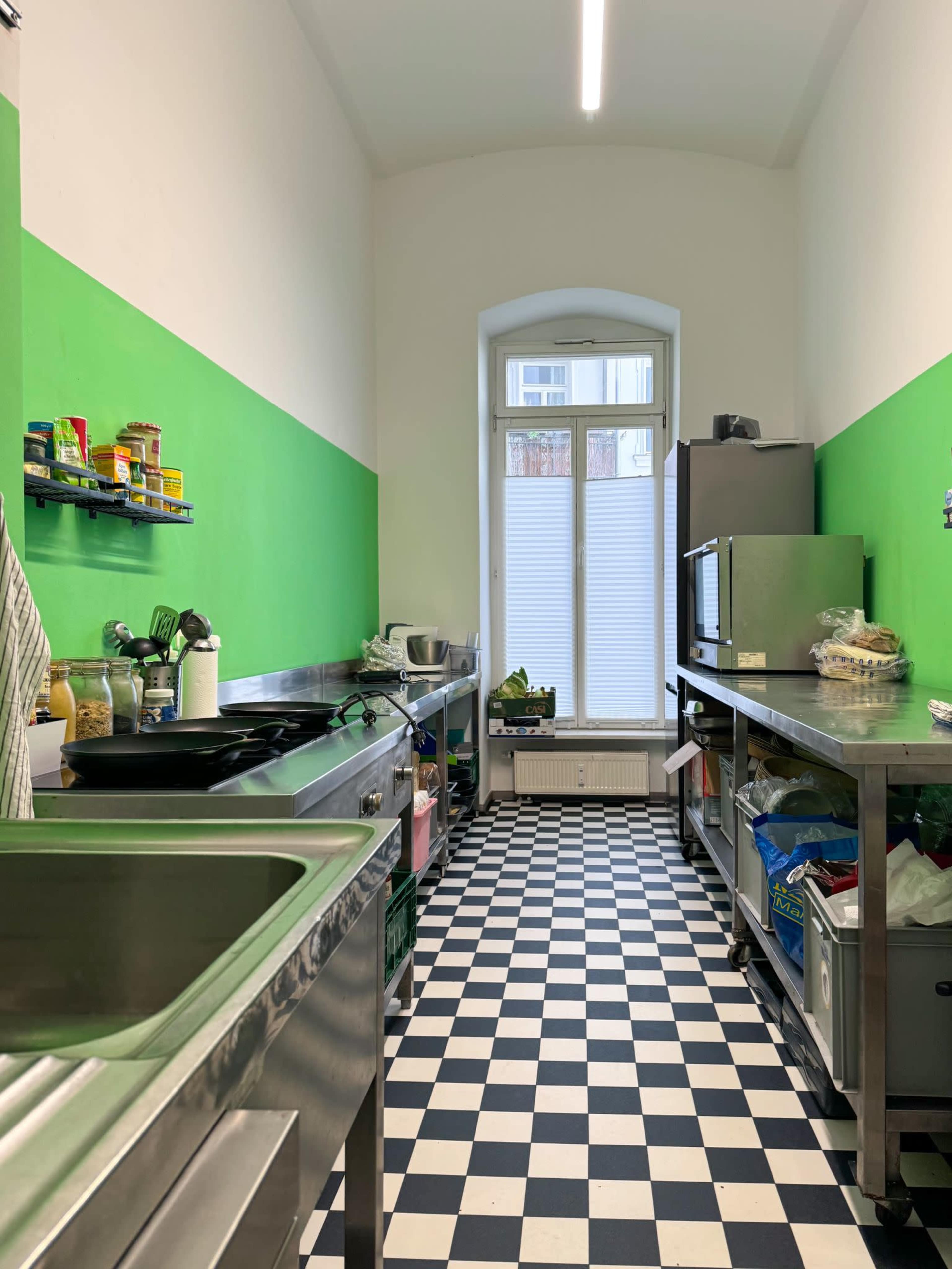 A narrow kitchen with green walls and a black-and-white checkered floor, featuring stainless steel counters and a window at the far end.