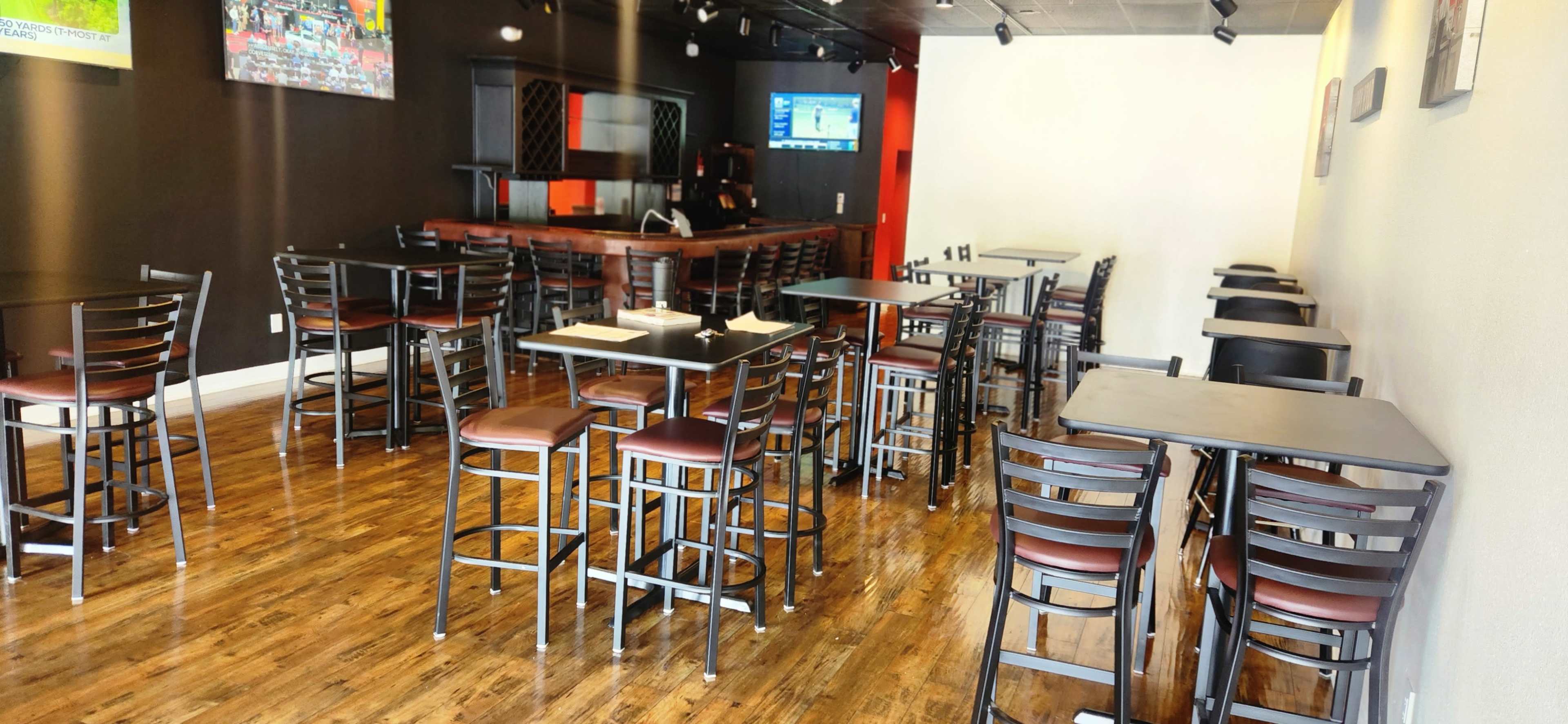 The image shows a partially empty restaurant with wooden floors, black and brown furniture, and a bar area in the background.