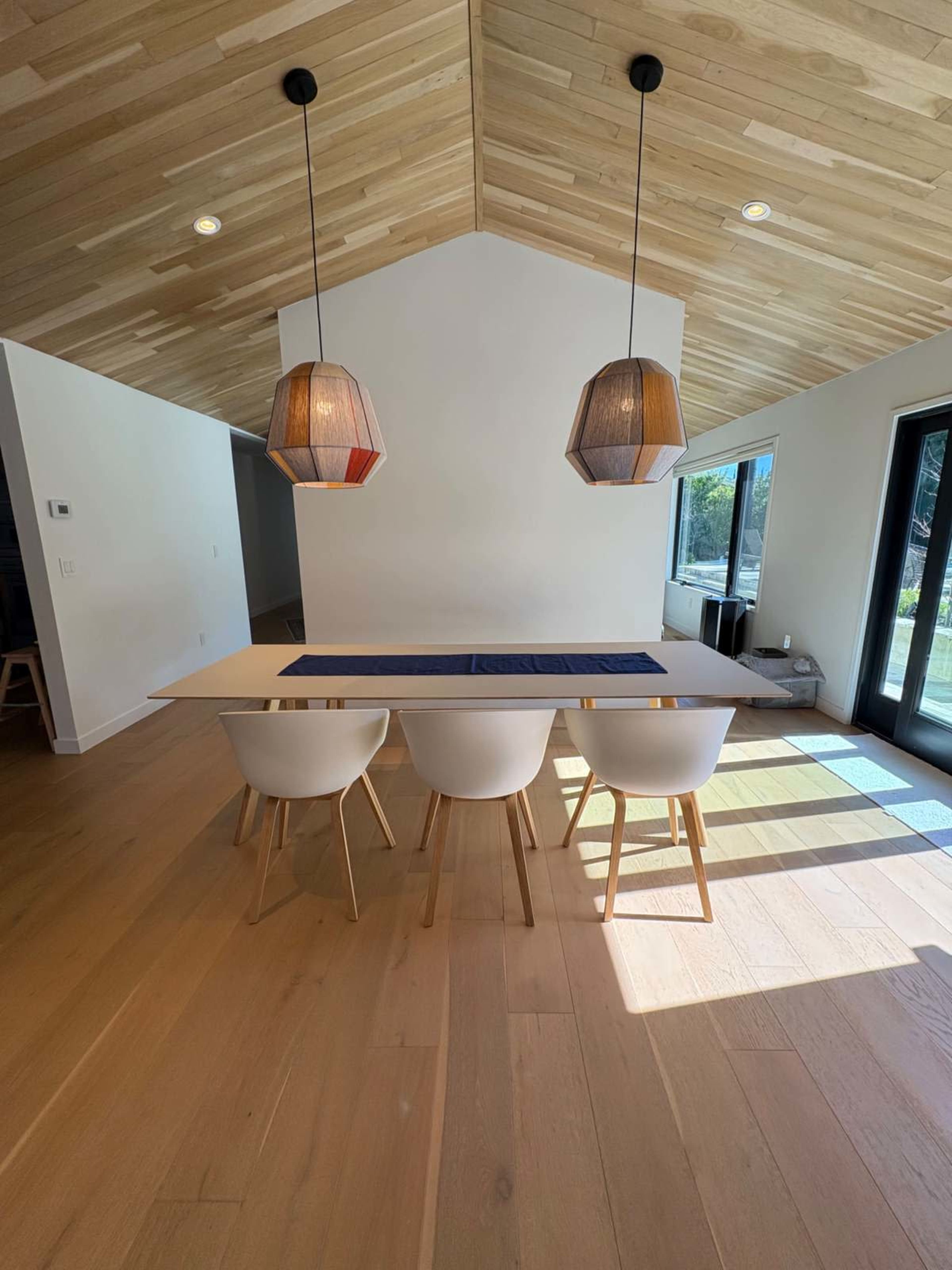 A dining area features a long table with four white chairs, illuminated by two pendant lights, under a sloped wooden ceiling.