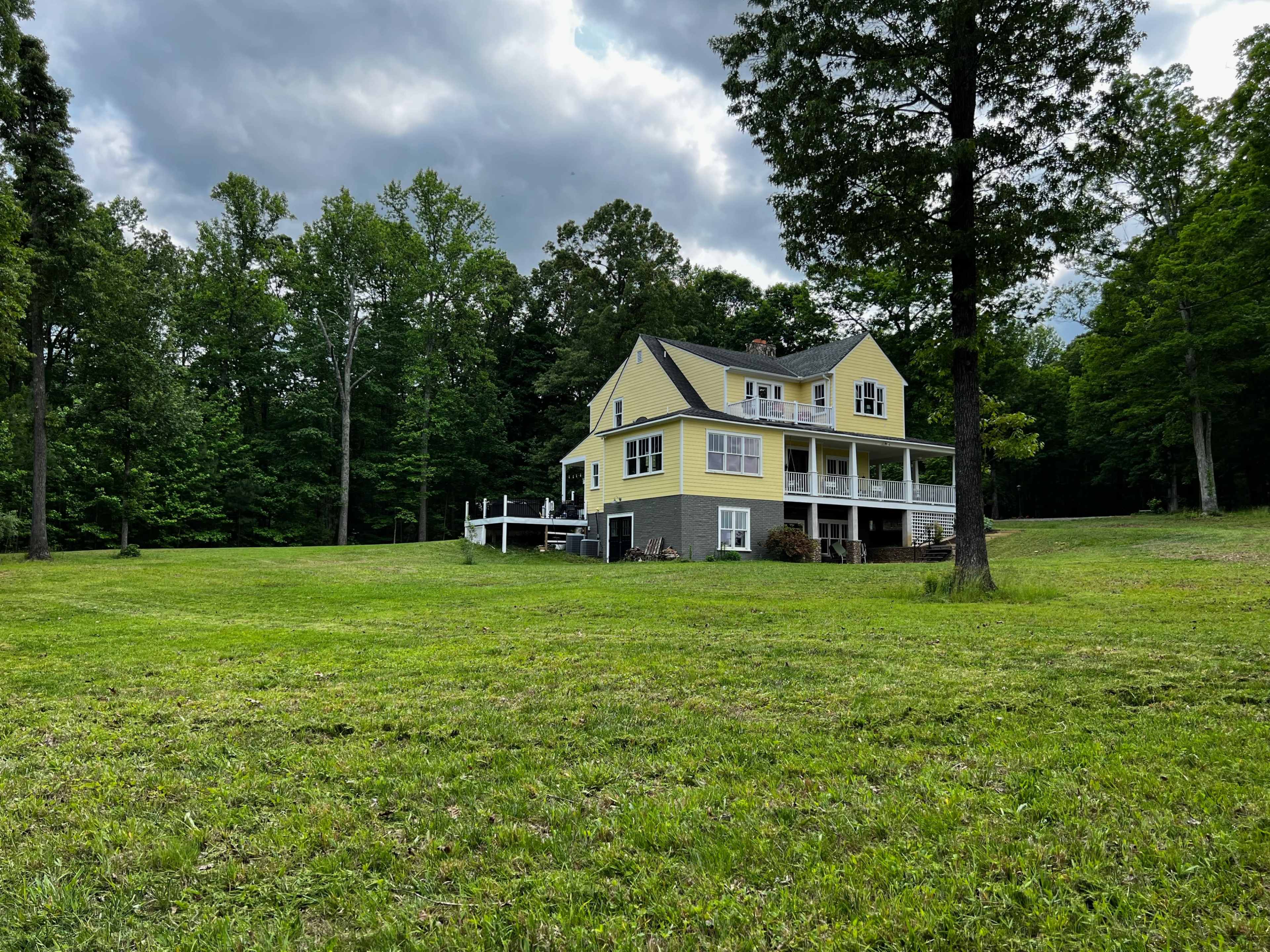 A two-story yellow house with a porch sits on a grassy lawn surrounded by green trees.