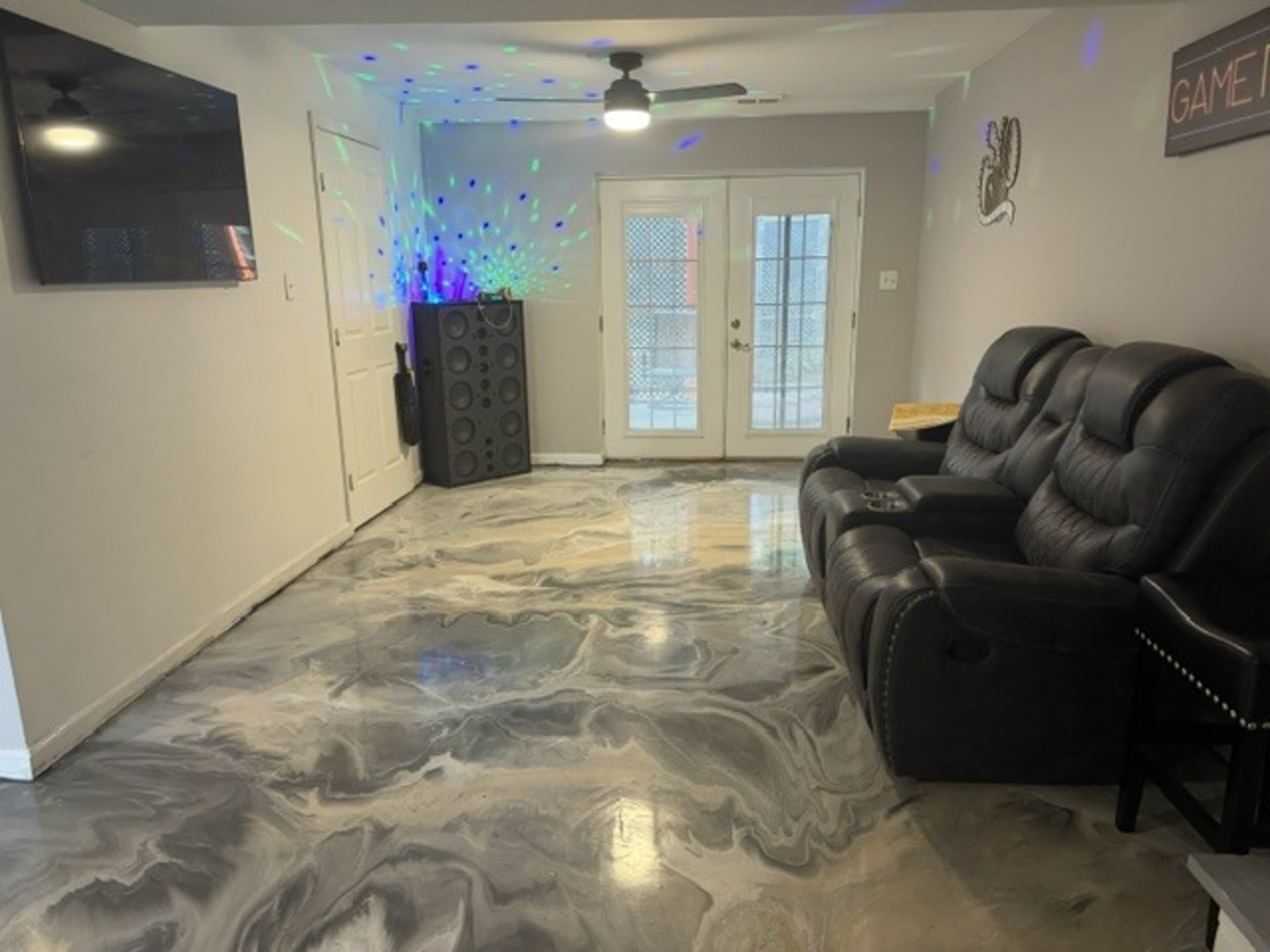 The image shows a room with a polished marble-like floor, a pair of black reclining chairs, a large speaker, and a door leading outside, illuminated by colorful lights.