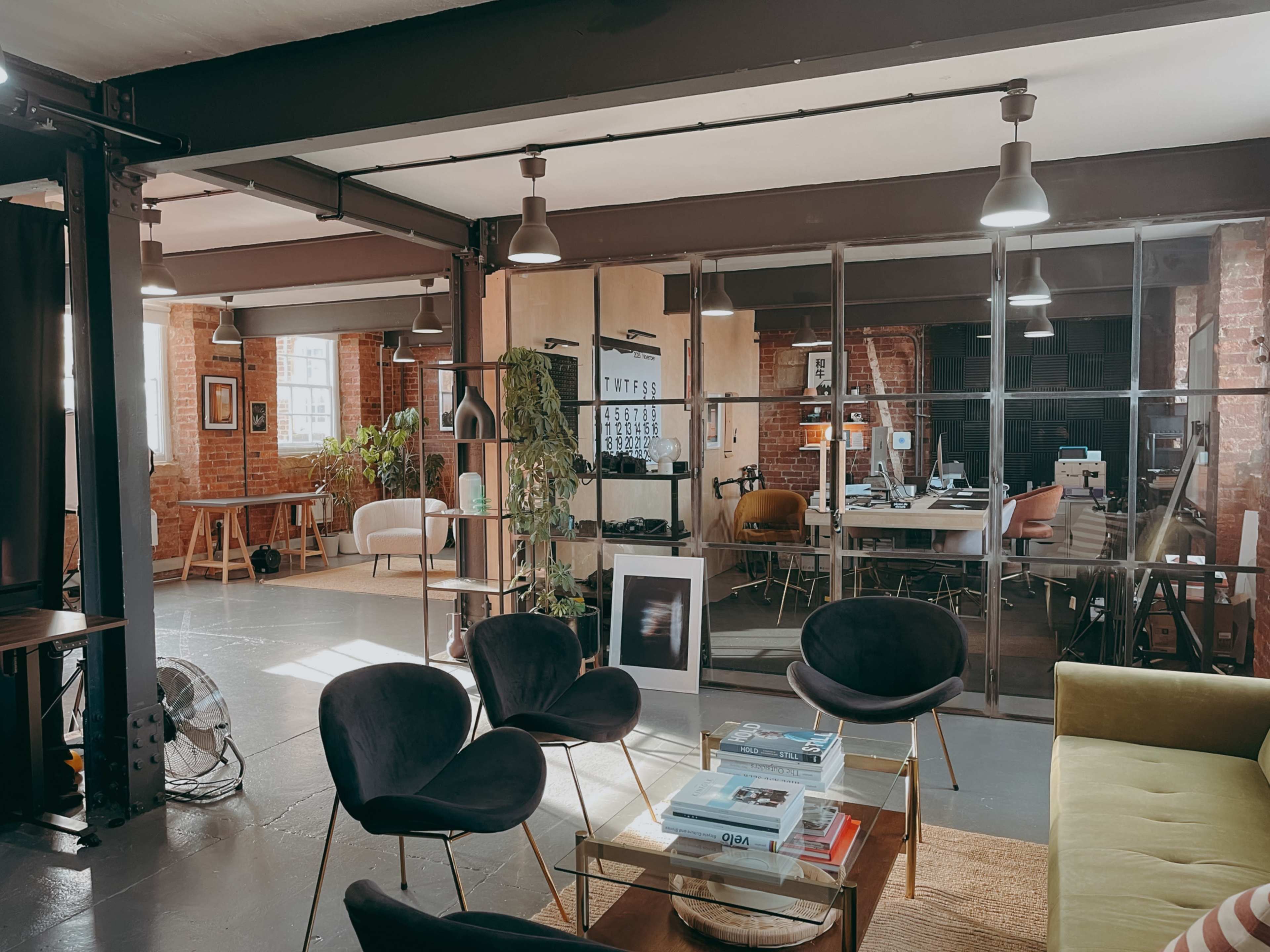 The image shows a modern office space featuring a lounge area with black chairs, a green couch, and large glass partitions separating workstations and meeting areas.