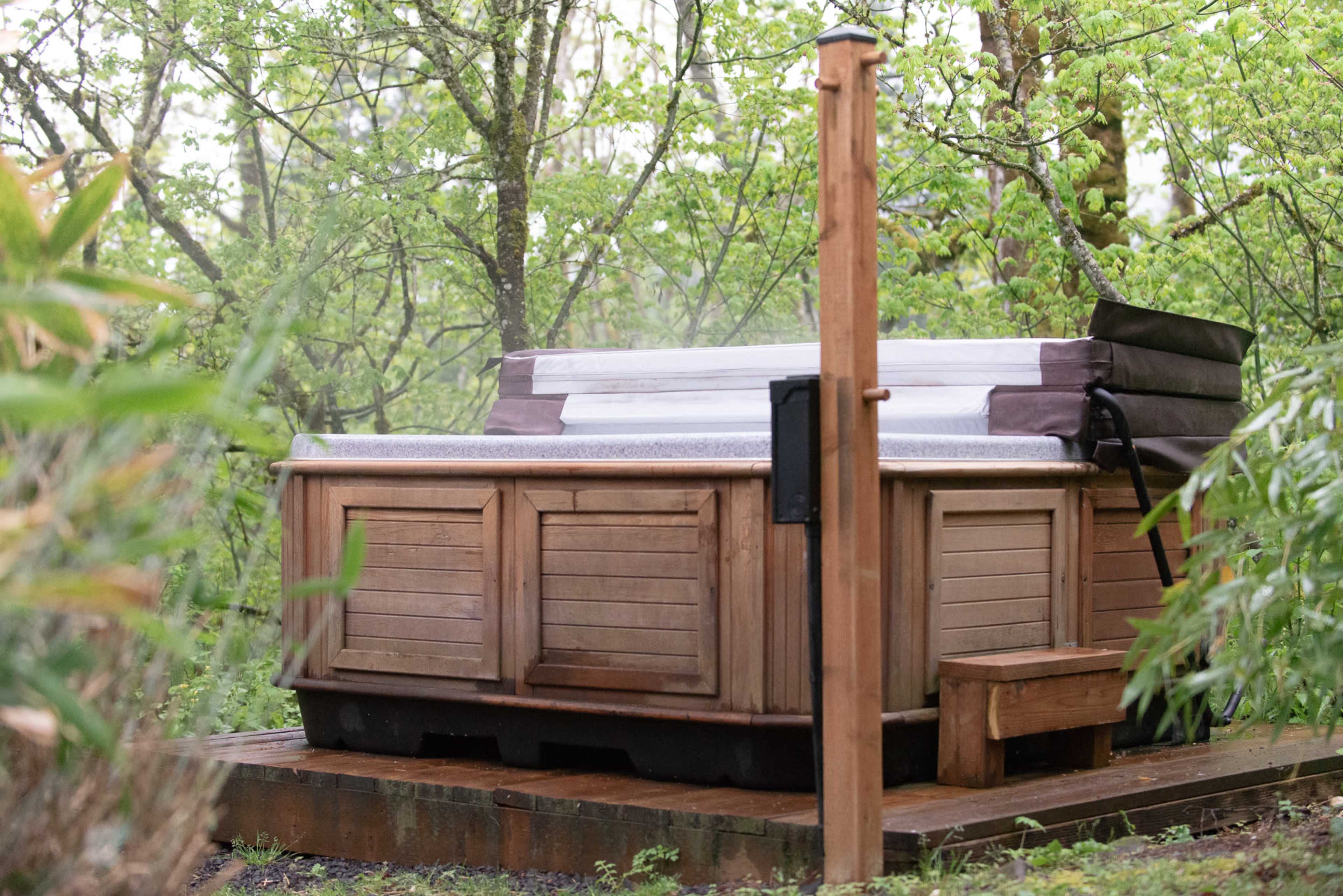 A wooden hot tub is situated on a wooden deck surrounded by lush greenery.