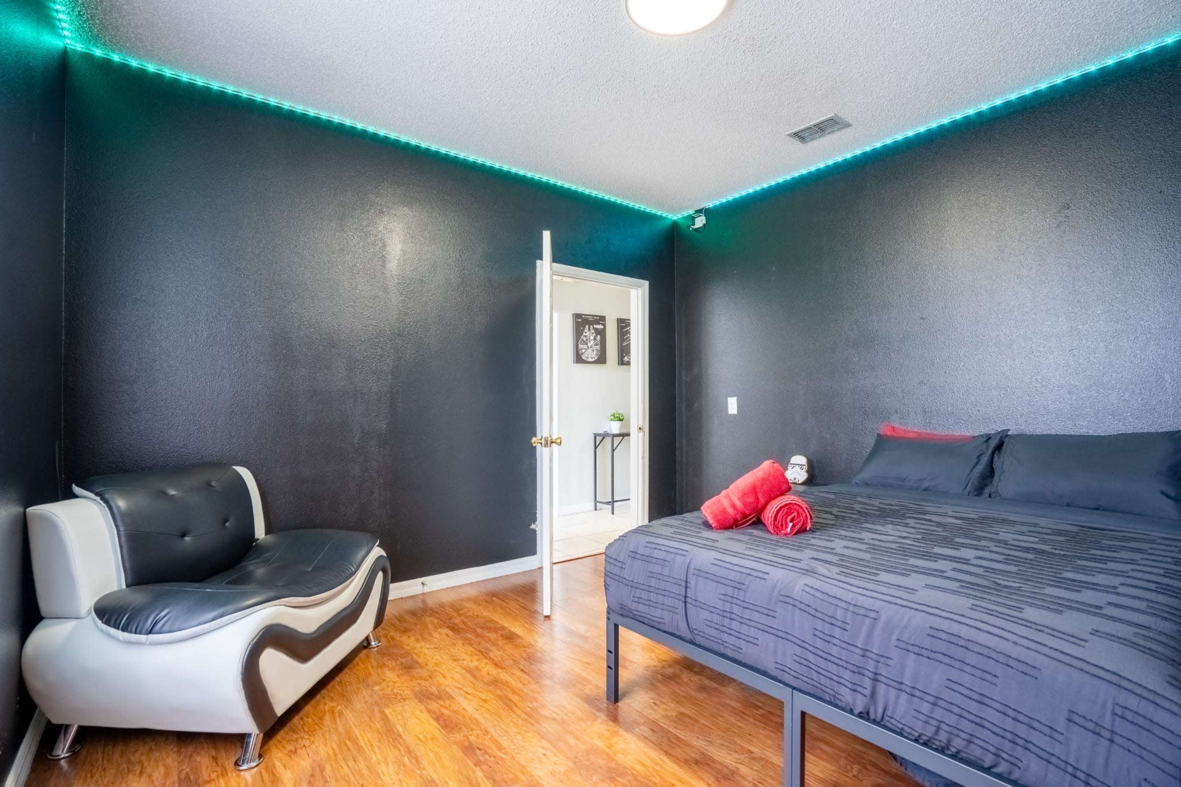 A bedroom with black walls, a bed made with dark bedding, and a gray chaise lounge in one corner.