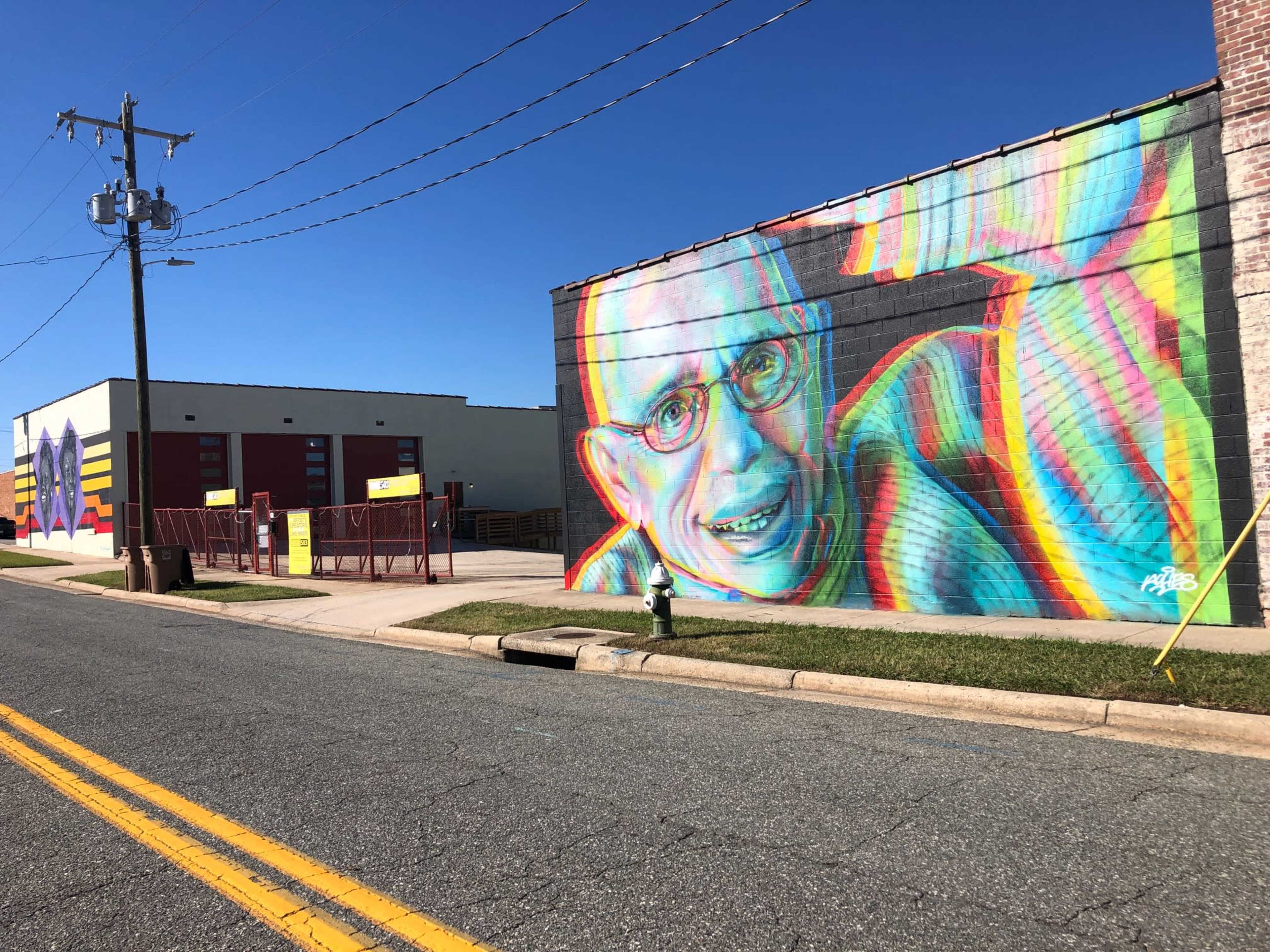 A colorful mural of a smiling man adorns the side of a brick building, with a power line and other commercial structures in the background.