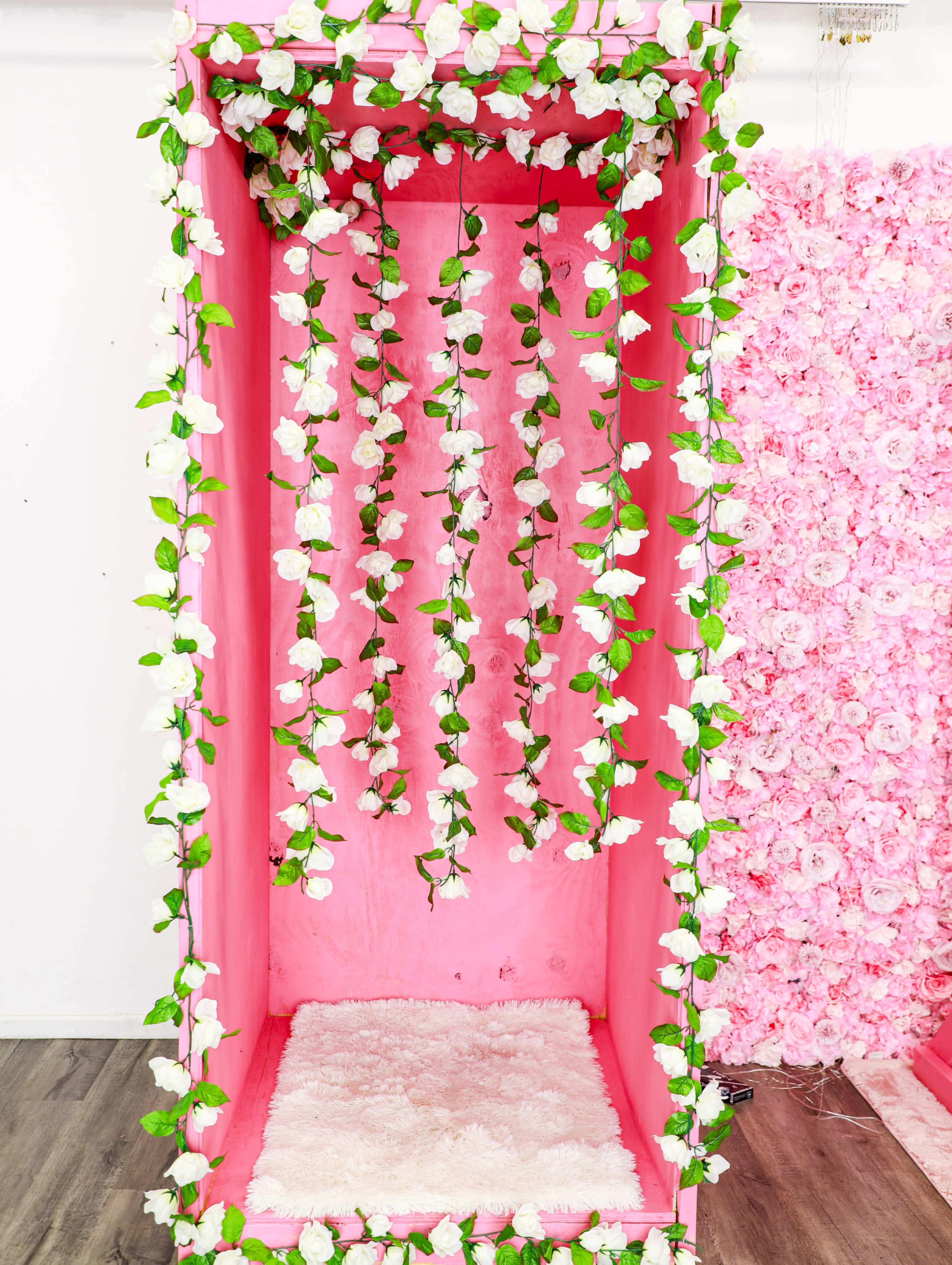 The image shows a pink photo booth decorated with white roses and greenery, featuring a fluffy white rug on the floor.