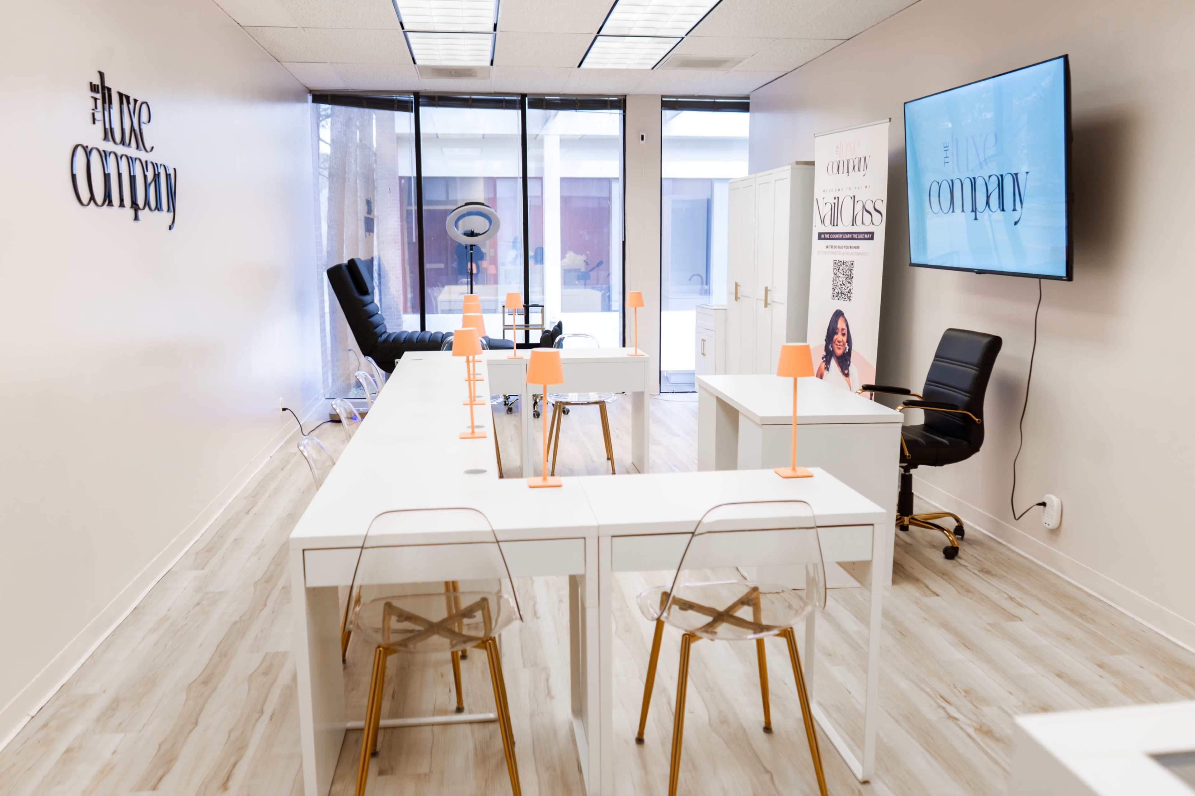 The image shows a modern workspace with multiple white desks, clear acrylic chairs, and a large screen displaying a logo, along with a stylish black chair and poster on the wall.