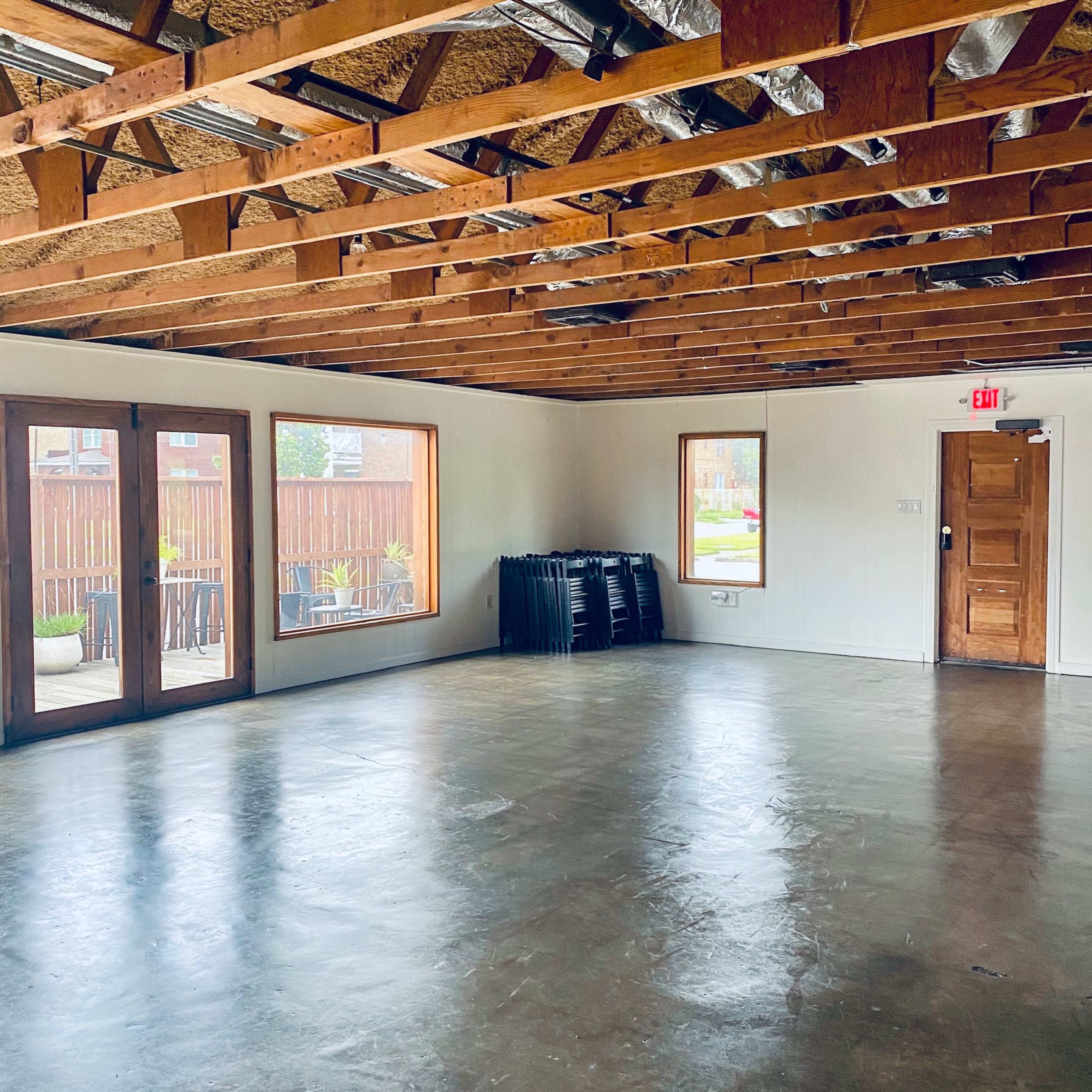 An empty room with exposed wooden beams, concrete flooring, and two large windows facing a patio.