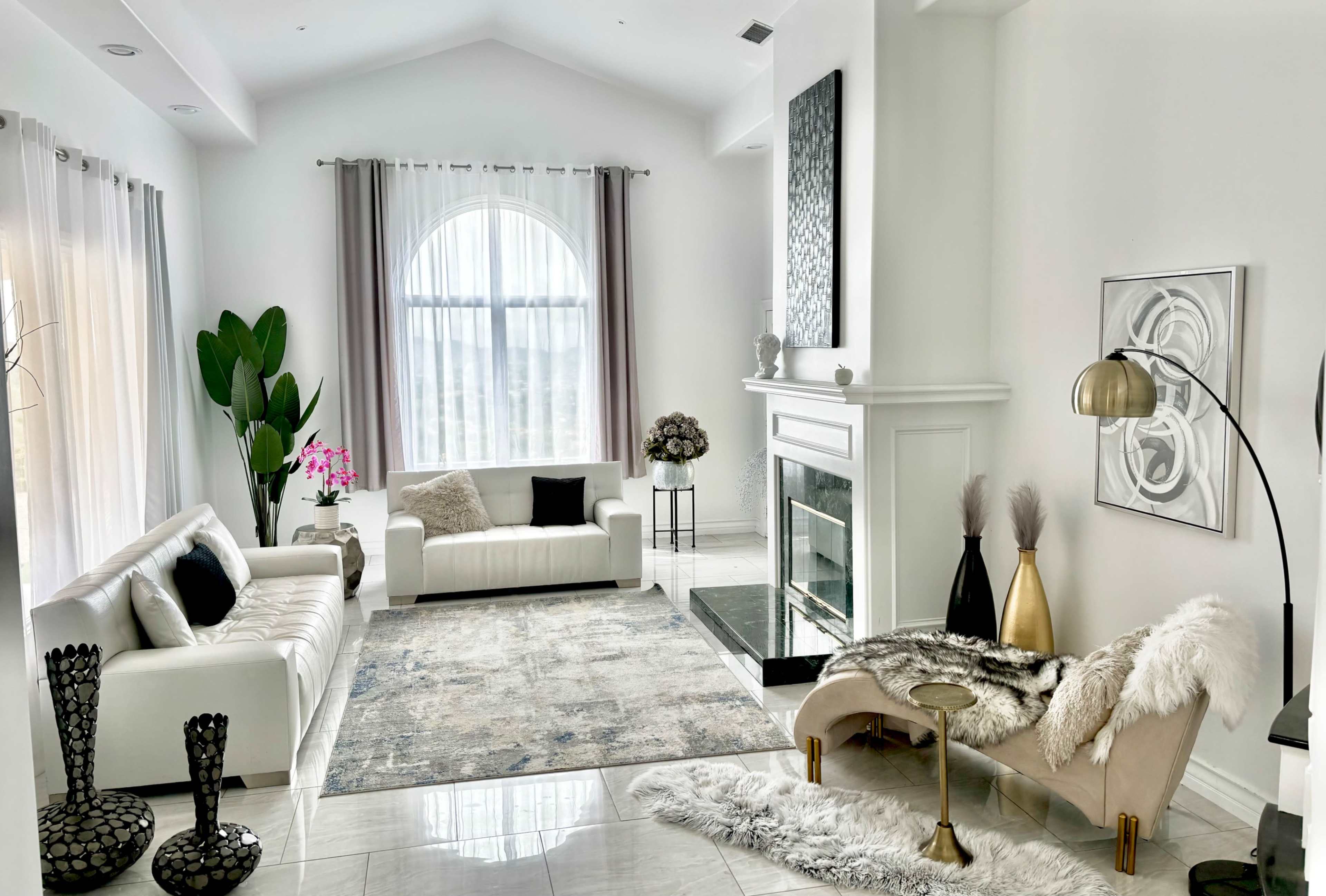 The image shows a modern living room with white sofas, a large window, a decorative fireplace, and a mix of plants and decor items.