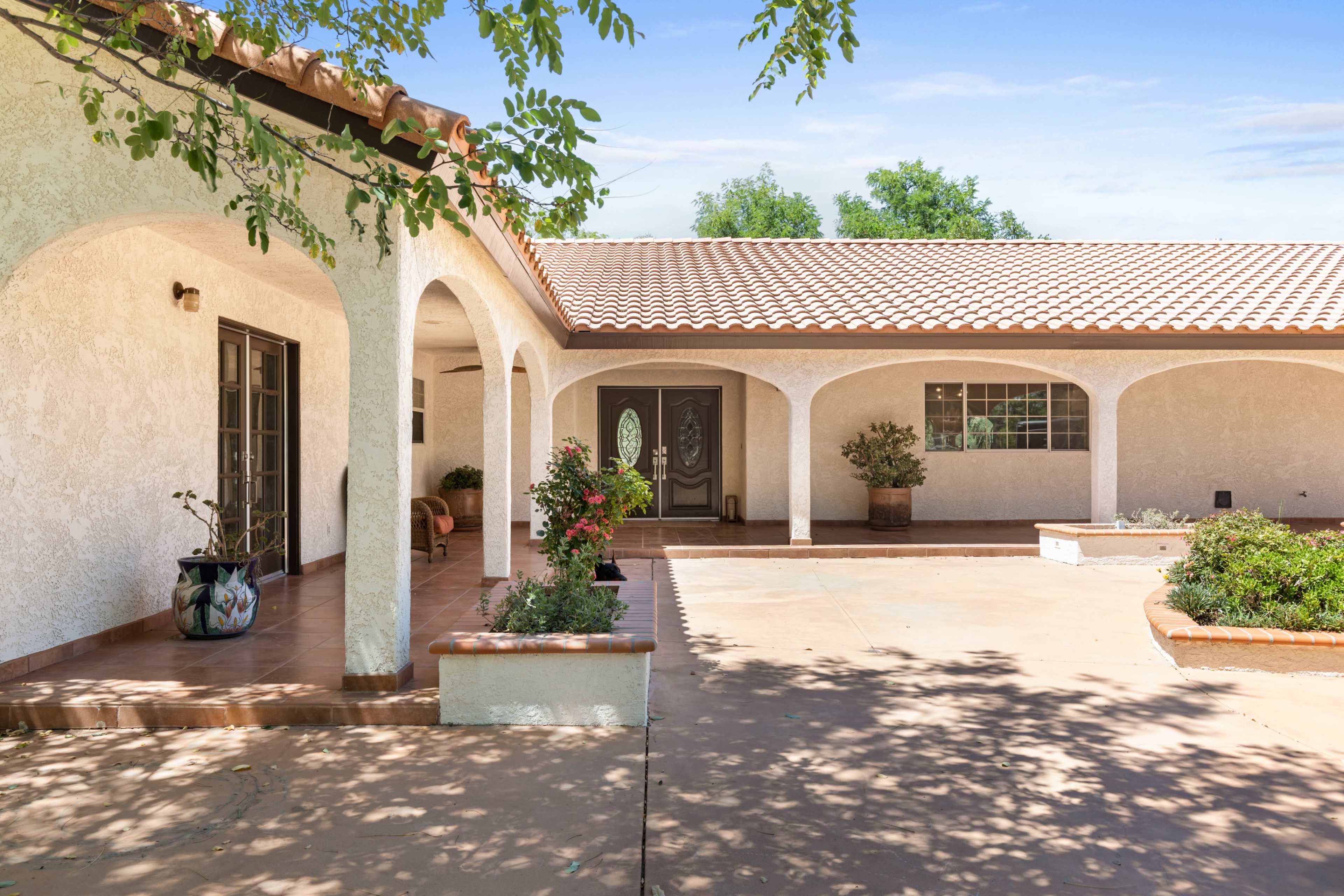 The image shows a single-story house with a tiled roof, arched doorways, and a courtyard featuring potted plants.