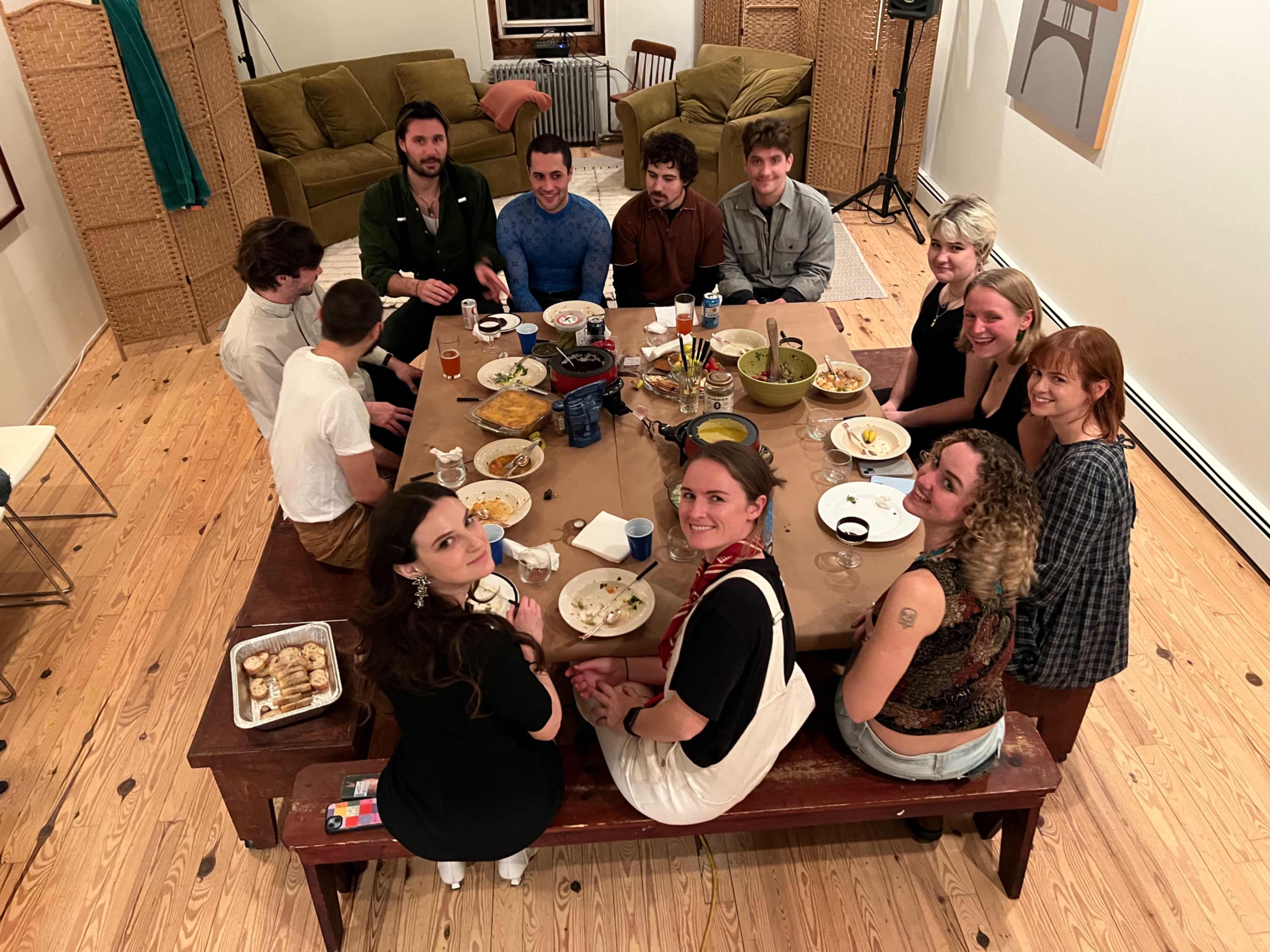 A group of twelve people sits around a large dining table filled with various dishes and drinks in a well-lit room.