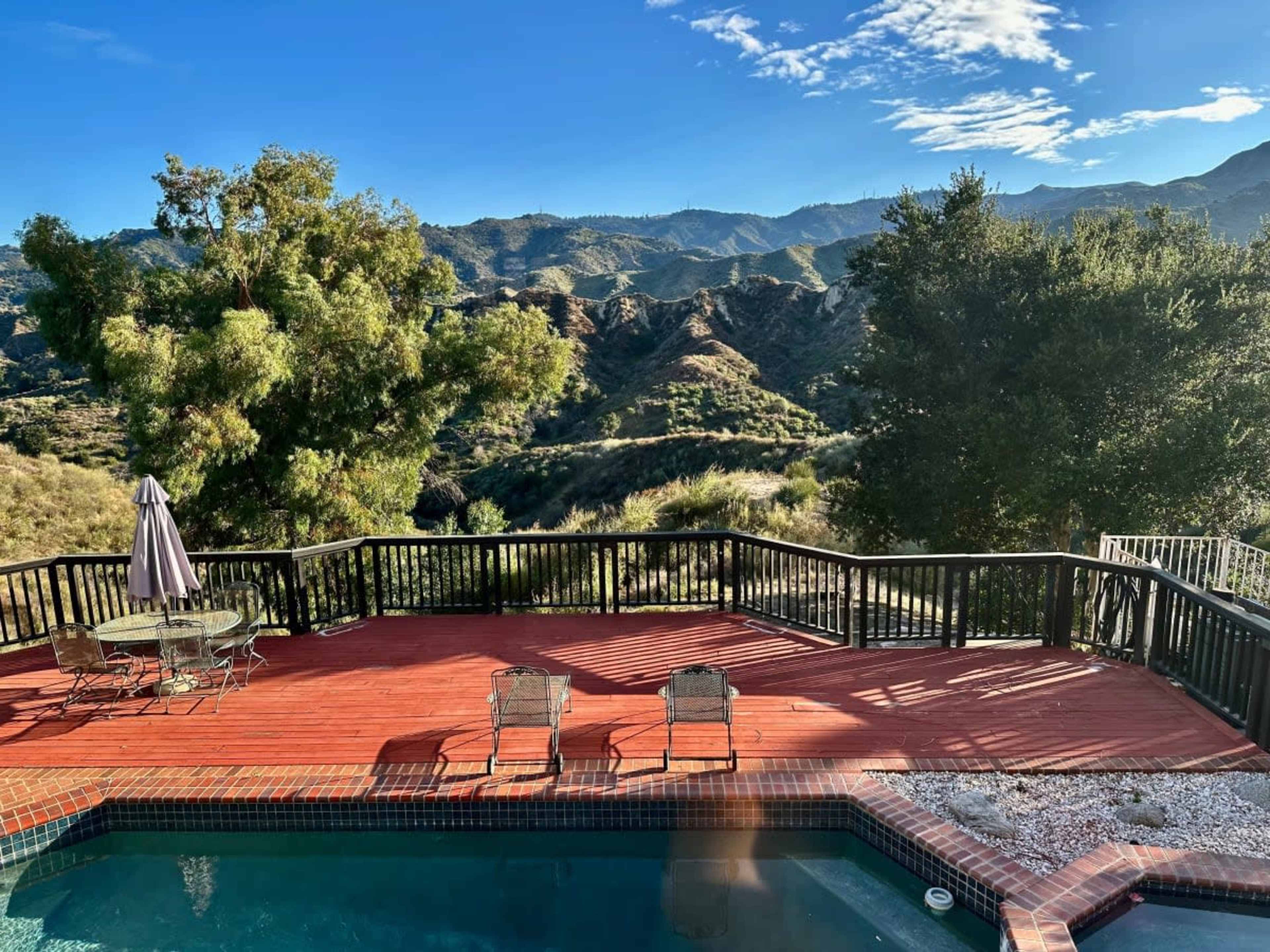 A wooden deck with two chairs and an umbrella overlooking a pool and mountainous landscape under a clear blue sky.