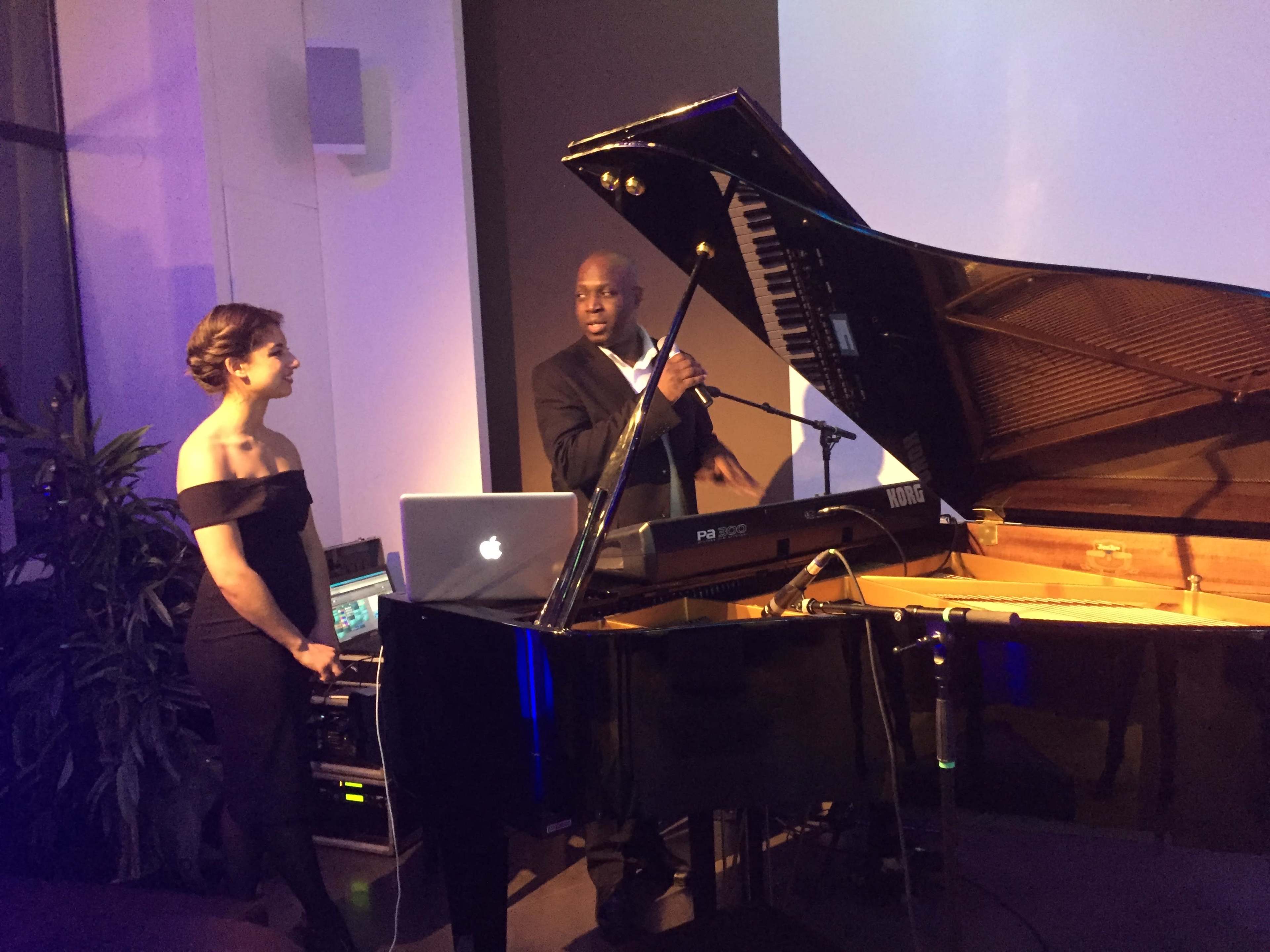 A man stands at a grand piano while a woman listens, as they prepare for a performance in a dimly lit setting.