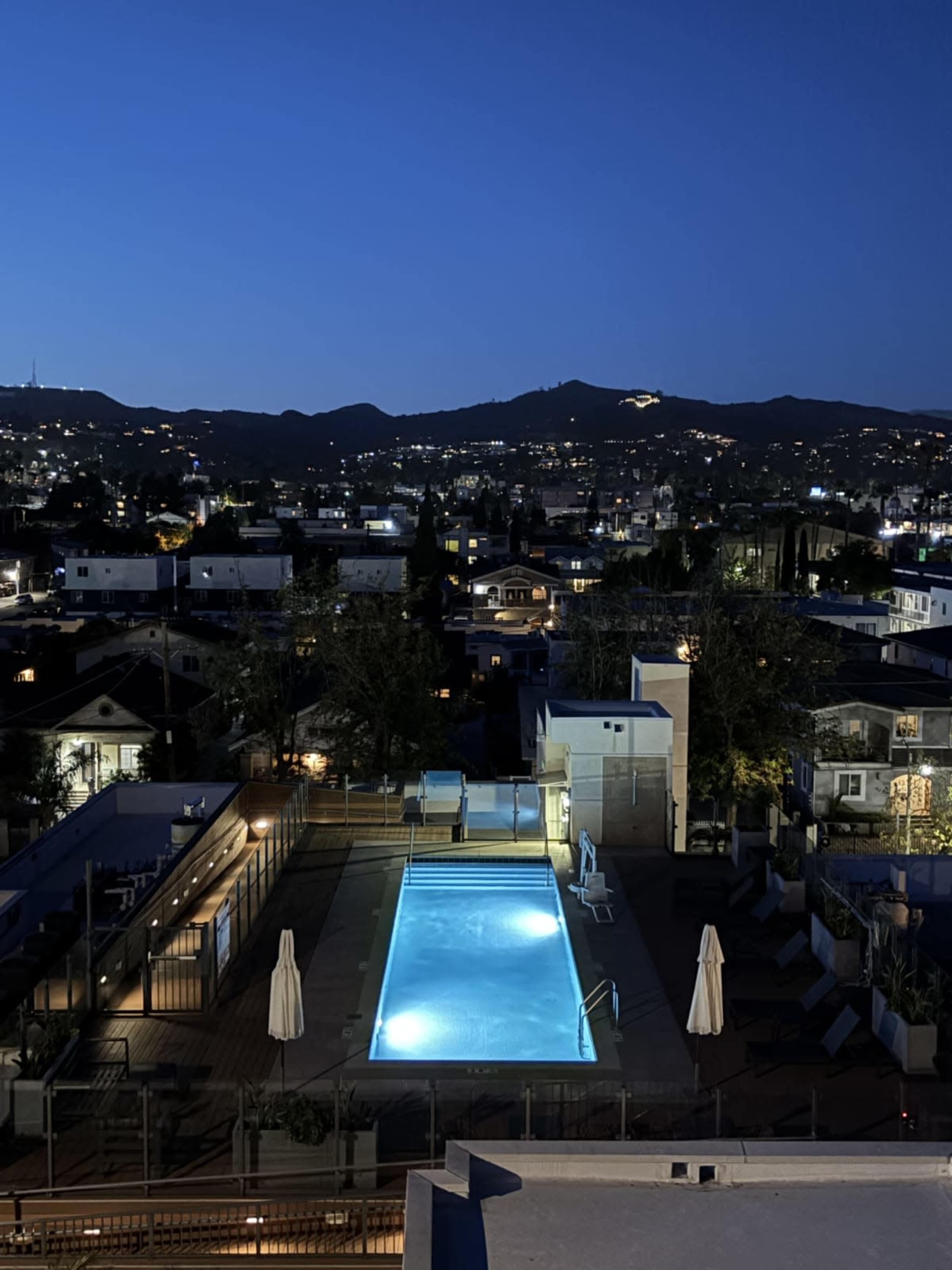 A well-lit swimming pool is visible on a rooftop terrace surrounded by city buildings, with mountains in the background at dusk.