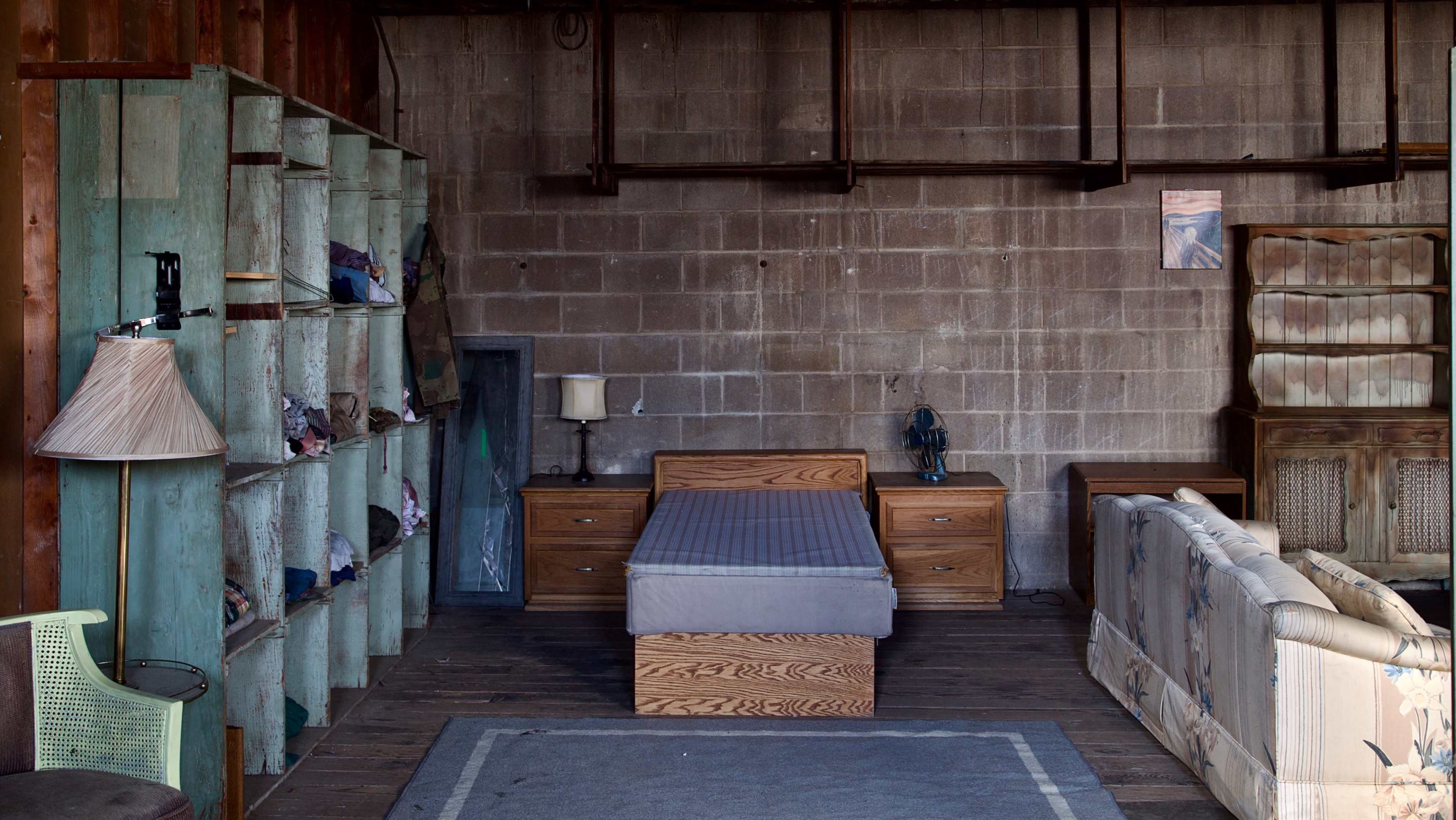 A sparsely furnished room features a wooden bed, a nightstand lamp, a vintage couch, and shelves filled with clothing against a gray cinderblock wall.