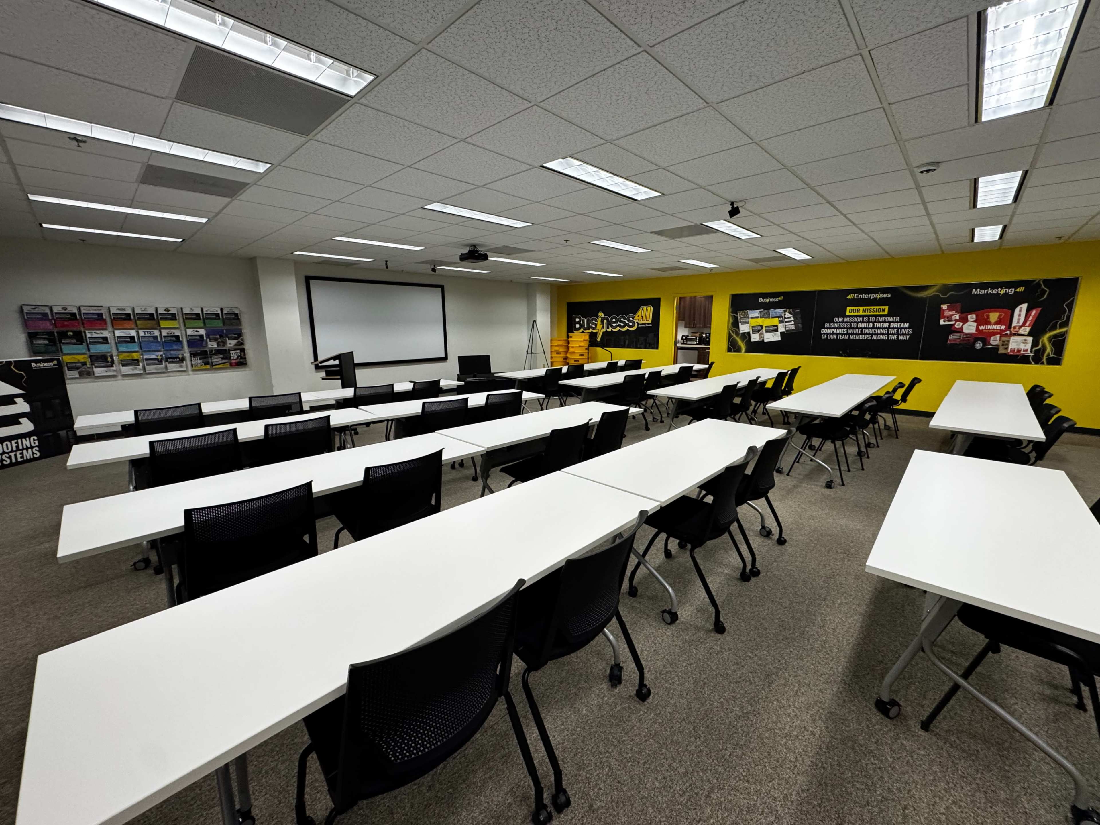 The image shows a neatly arranged classroom with multiple rows of white tables and black chairs, complemented by a projector screen and yellow wall graphics.