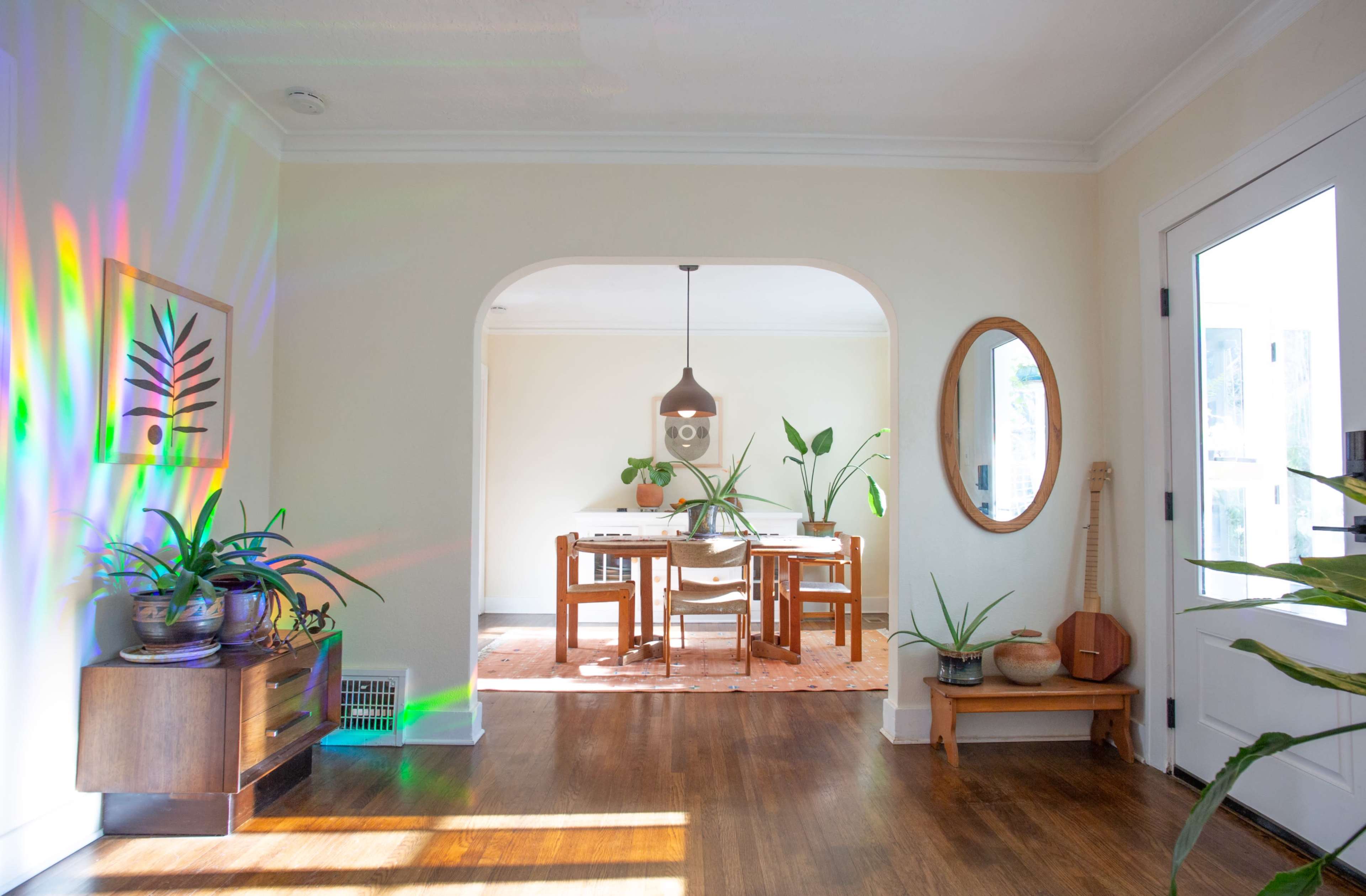 A brightly lit entryway leads to a dining area adorned with plants and a wooden table, featuring a mirror on one wall and a guitar in the corner.