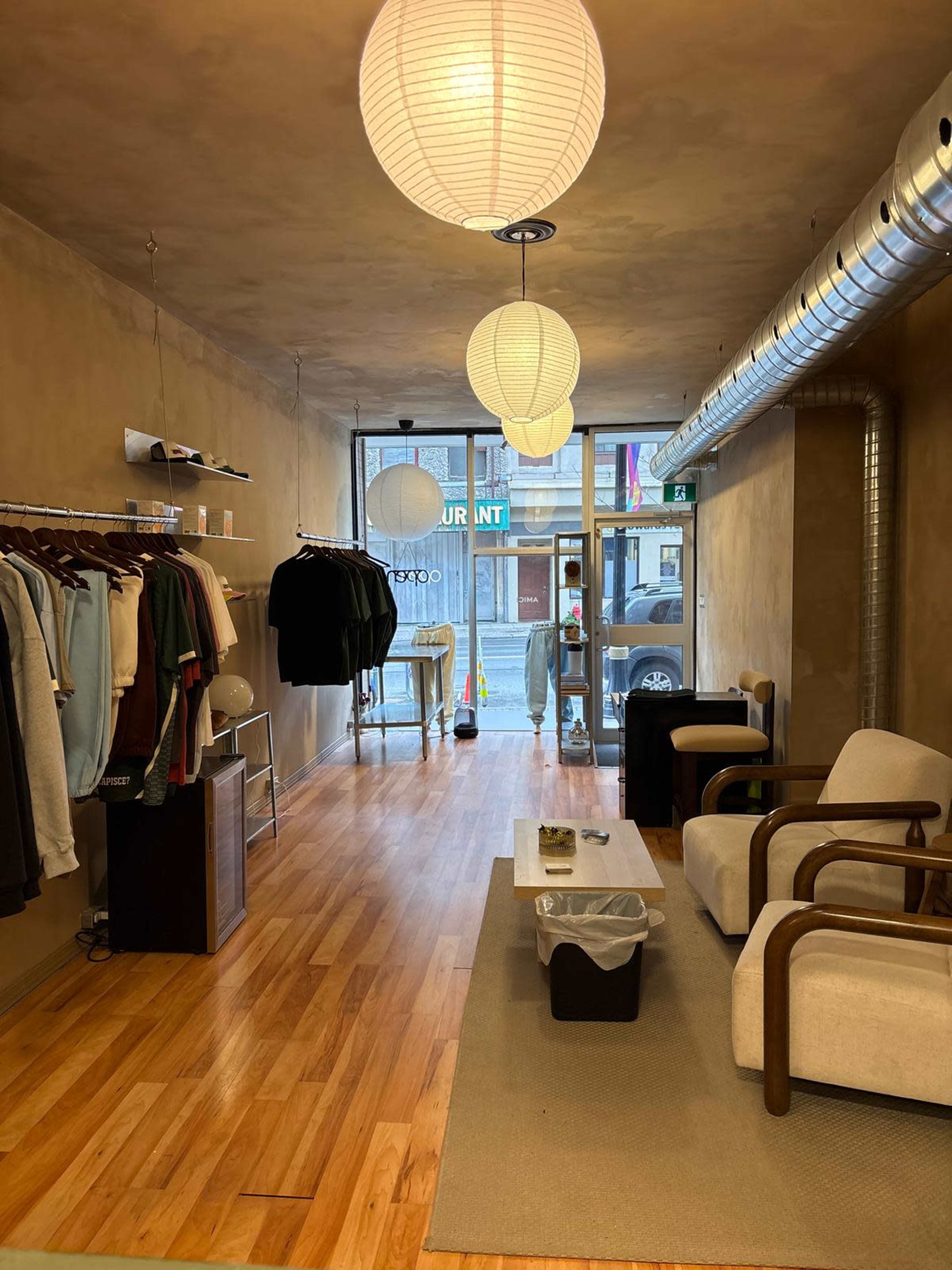 The image shows a minimalist retail space with wooden flooring, hanging clothing racks, a seating area, and large windows at the front displaying a street view.