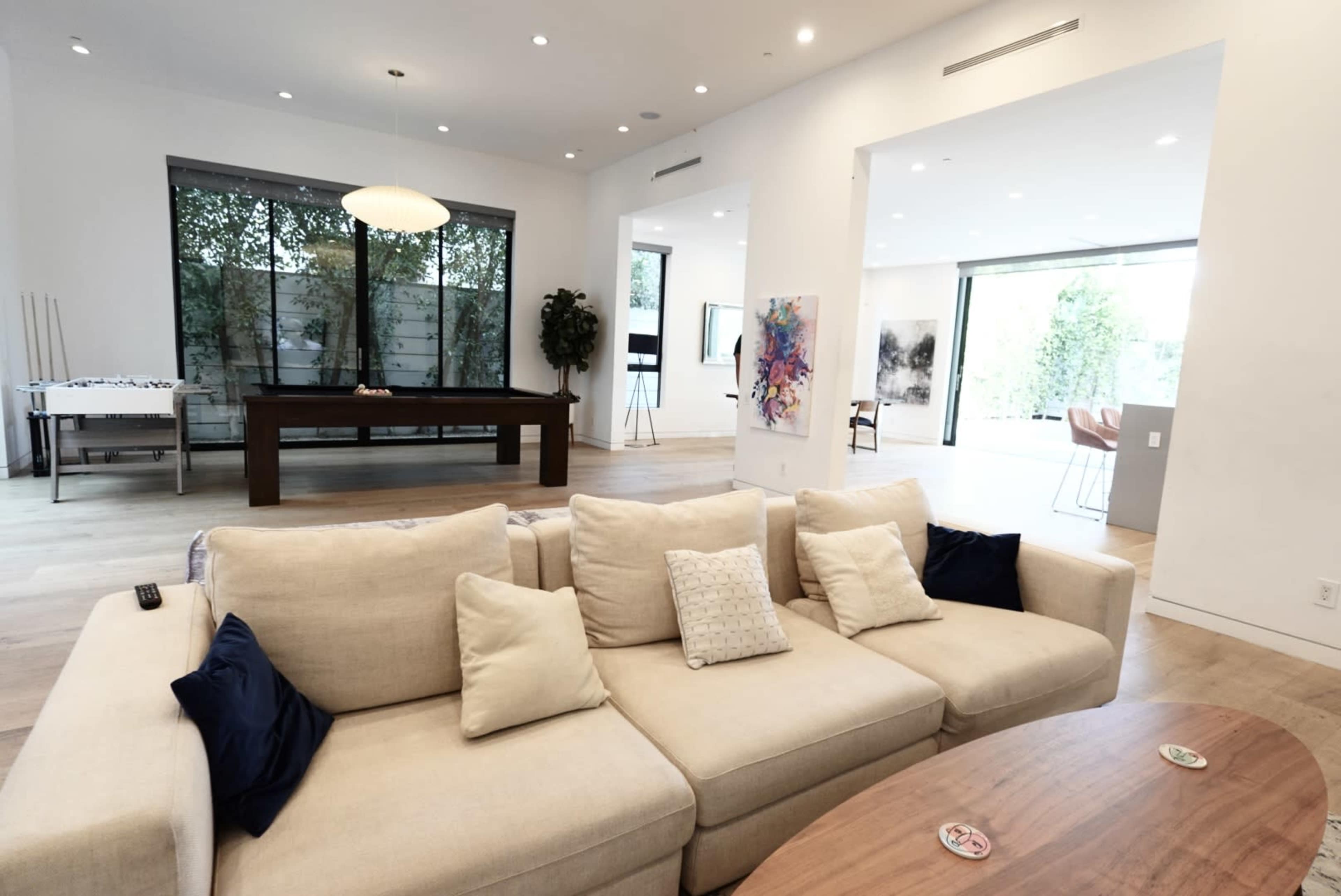 The image shows a spacious living room featuring a beige sectional sofa, a wooden coffee table, and large windows that provide natural light.